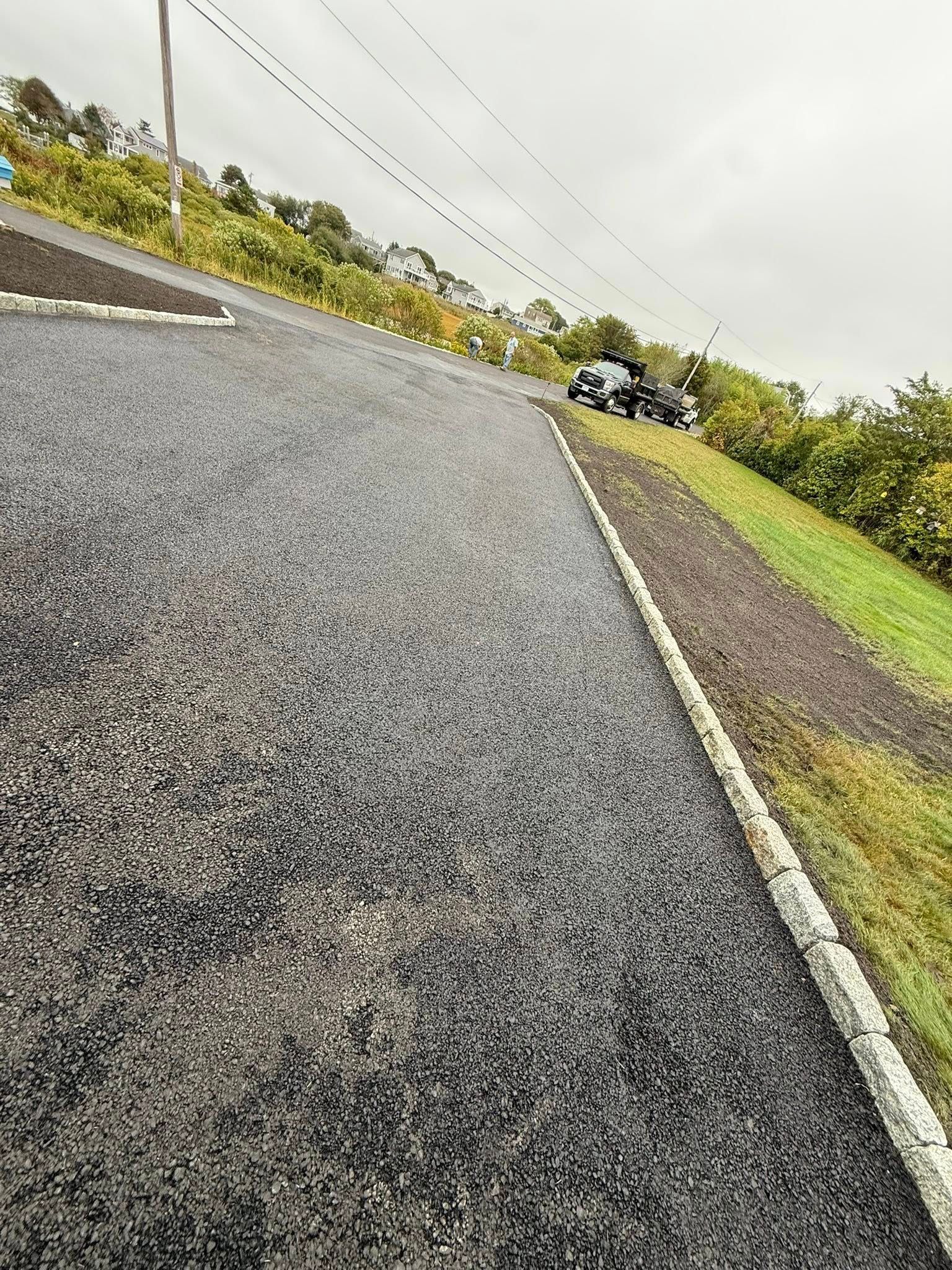 A freshly paved gray asphalt driveway with a stone border, angled upward toward a grassy hill under a cloudy sky.