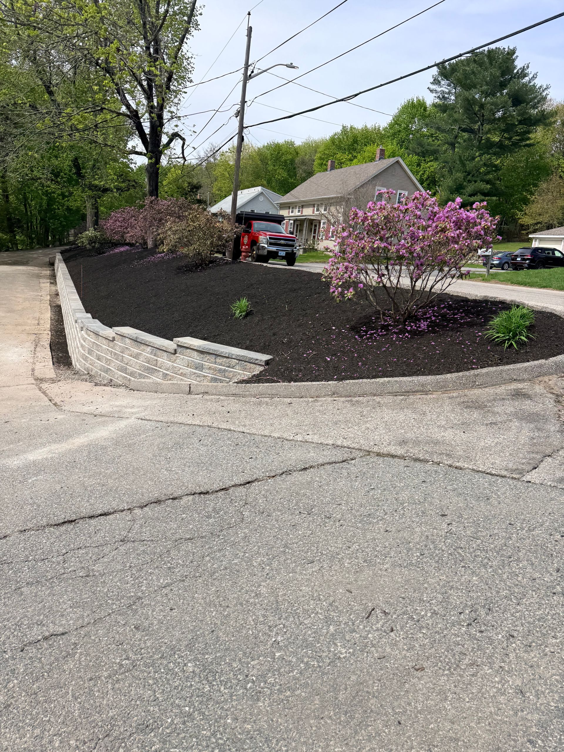 Curved flower bed with pink flowering shrubs beside a driveway and house under a bright sky
