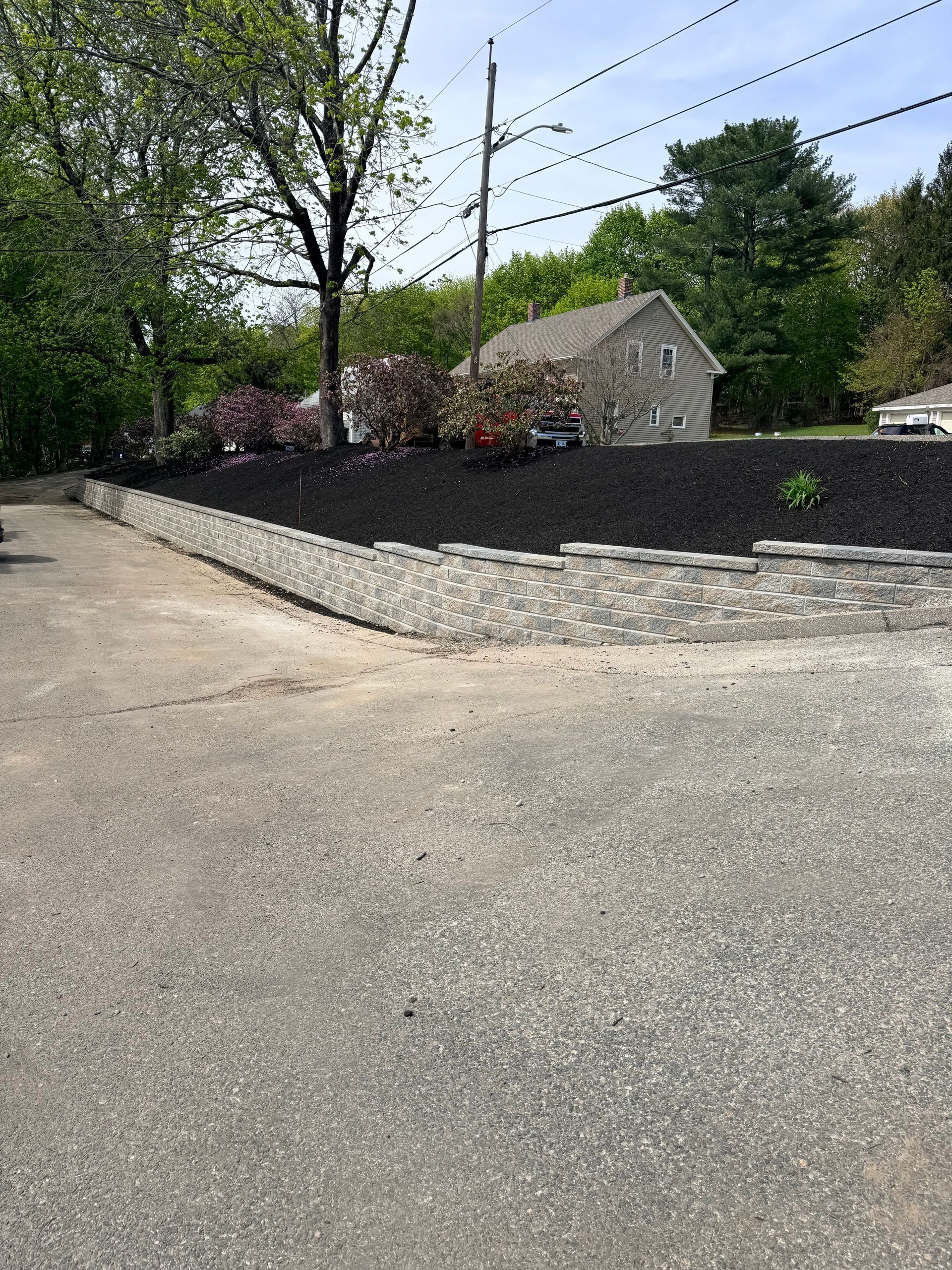 Stone retaining wall beside a paved road, with trees and a flower bed on a sunny day