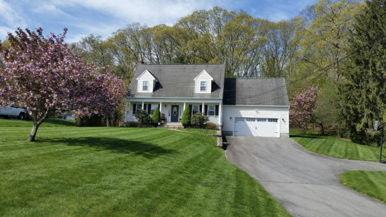 White house with a gray roof, dormers, and an attached garage. Lush green lawn, driveway, and blooming pink trees in a bright, sunny setting.