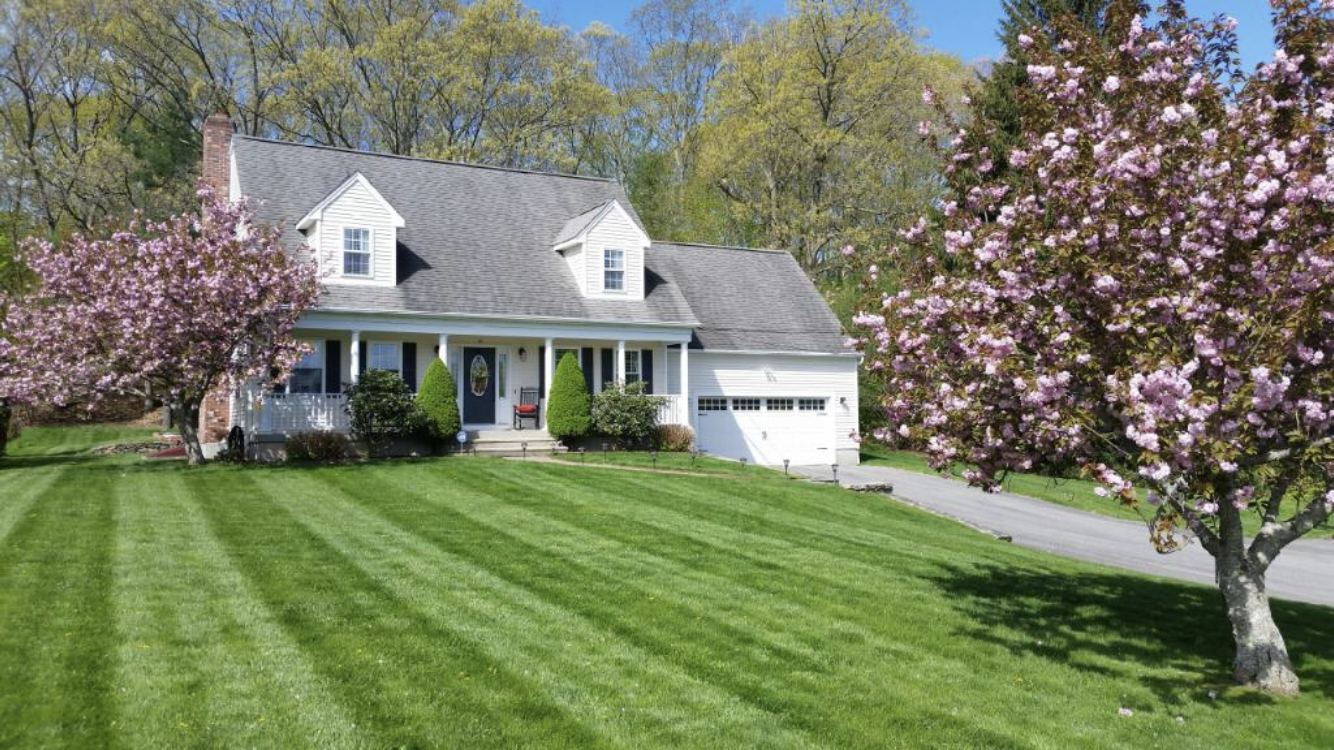 A well-manicured lawn with striped patterns leads to a white house, framed by blossoming pink trees, on a sunny day.