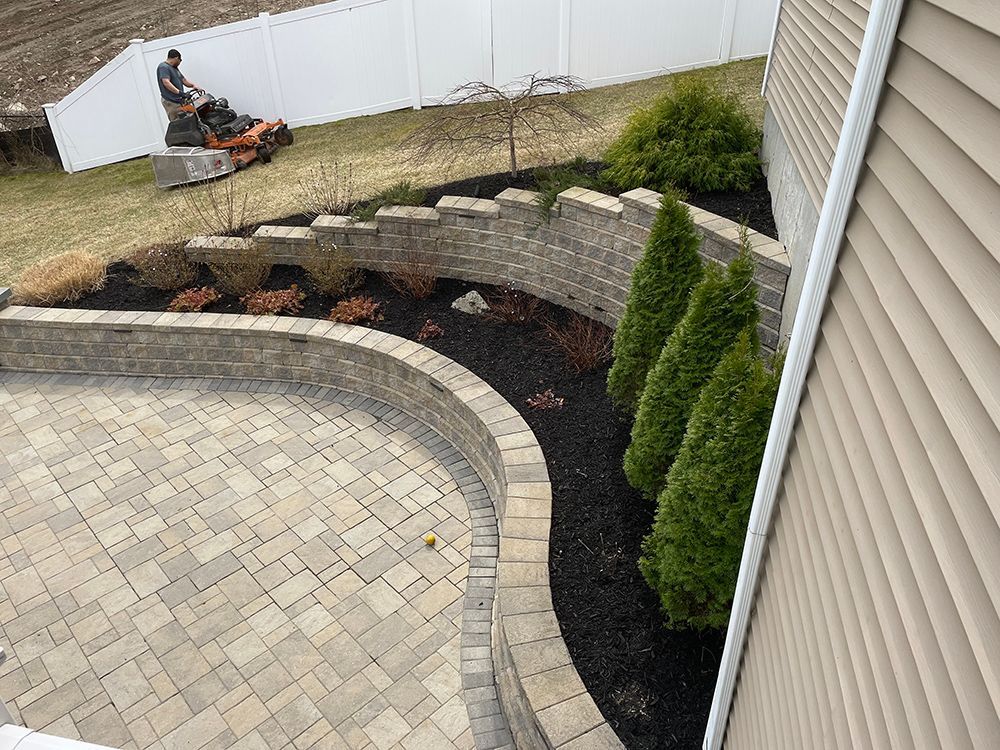 A man is riding a lawn mower on a patio next to a house.