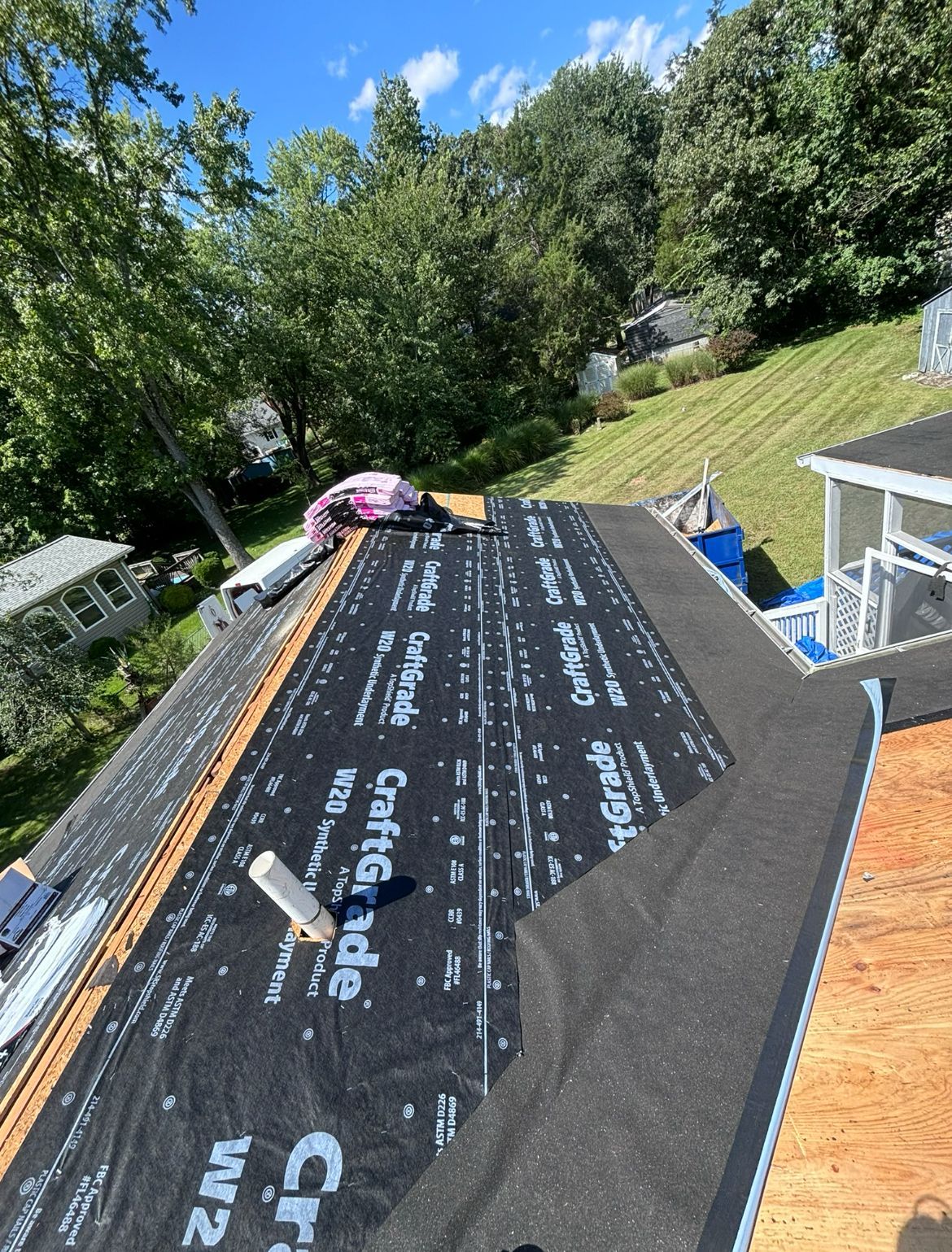 Roof under construction with black underlayment and plywood, surrounded by trees under a sunny sky.