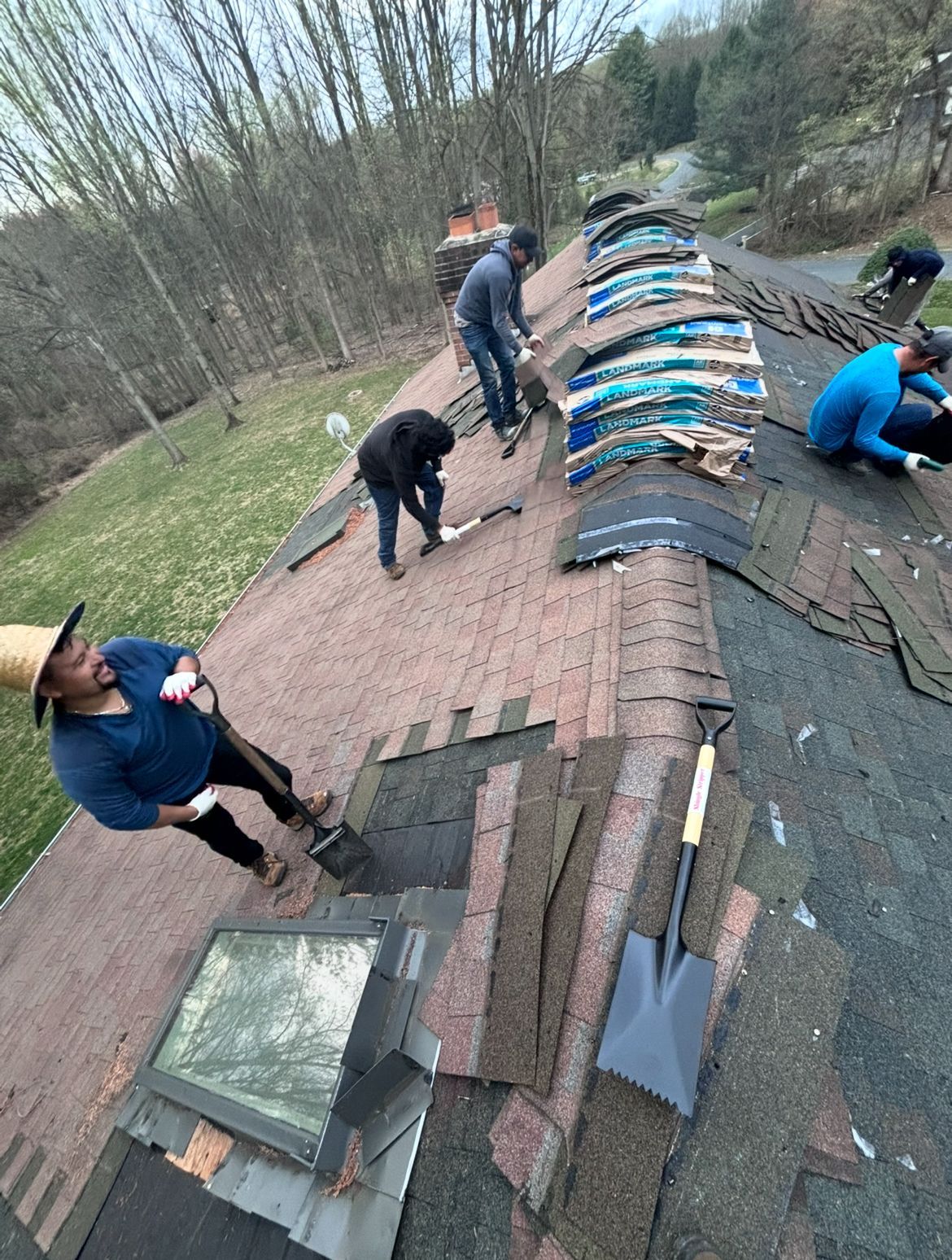 Roofers working on a house roof, removing old shingles. A person pulls shingles near a skylight.