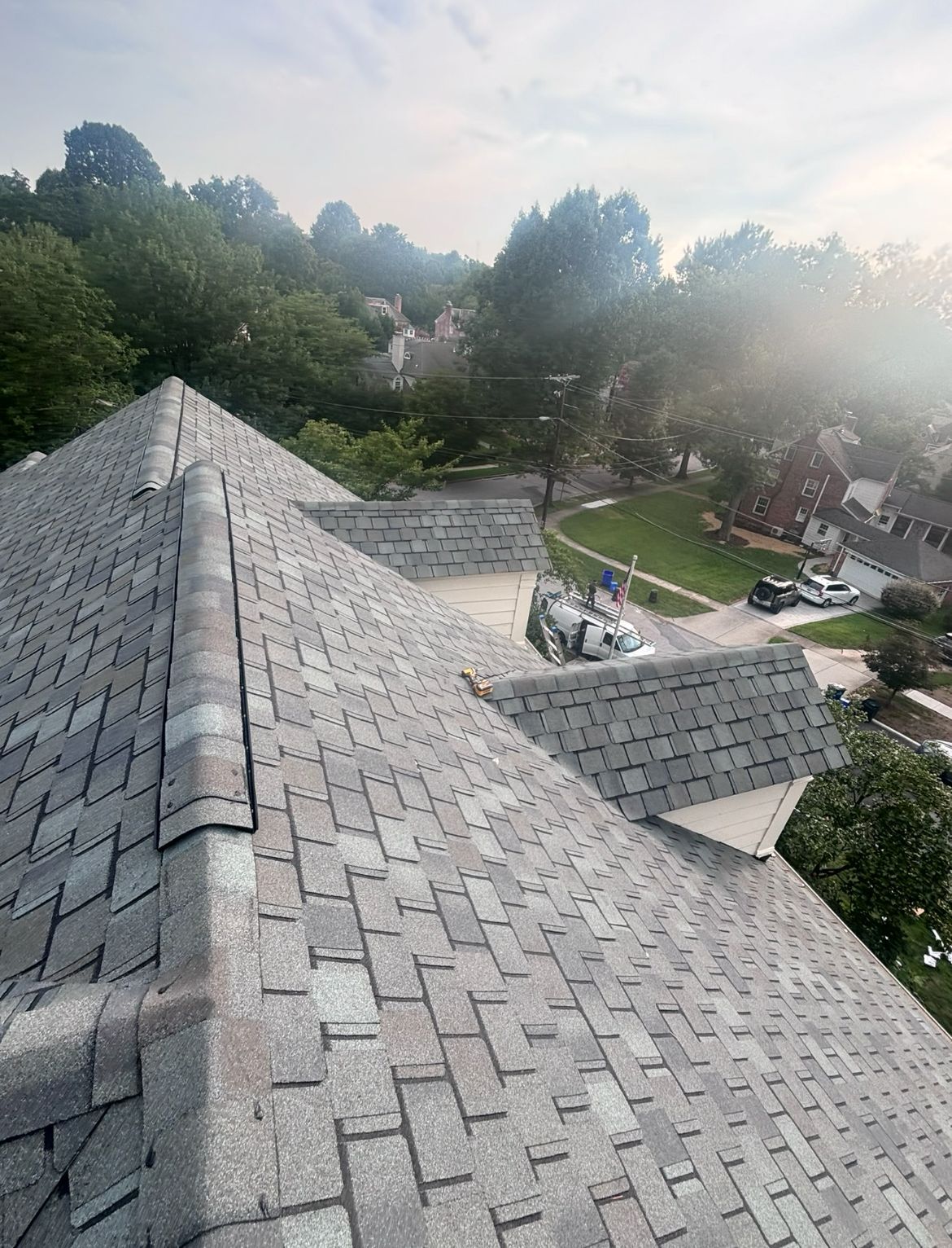 Gray asphalt shingle roof on a house, with trees and a street in the background on a sunny day.