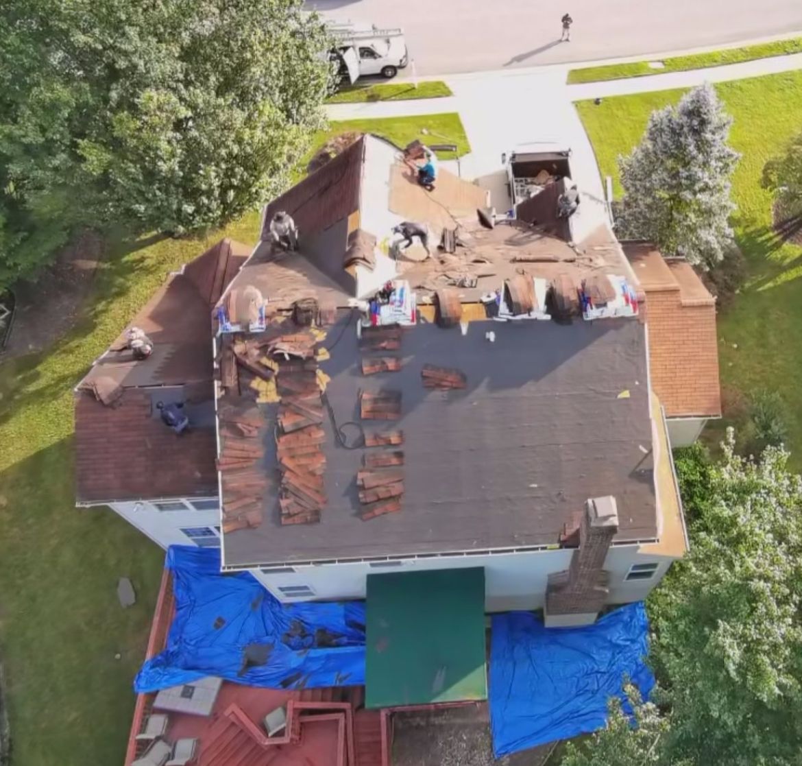 Roofers replacing a roof on a two-story house. Blue tarps cover the lower level. Trees surround the house.