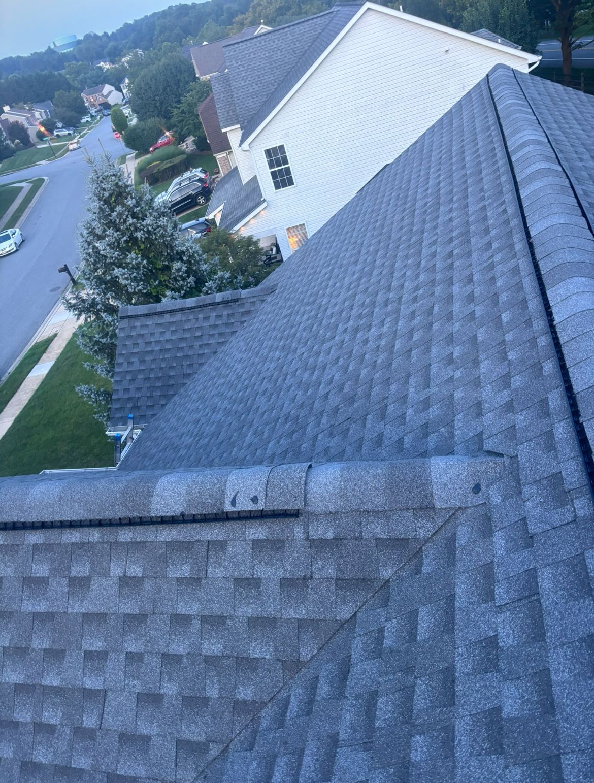 View from a rooftop shows gray asphalt shingles. Houses, trees, and a street are in the background.