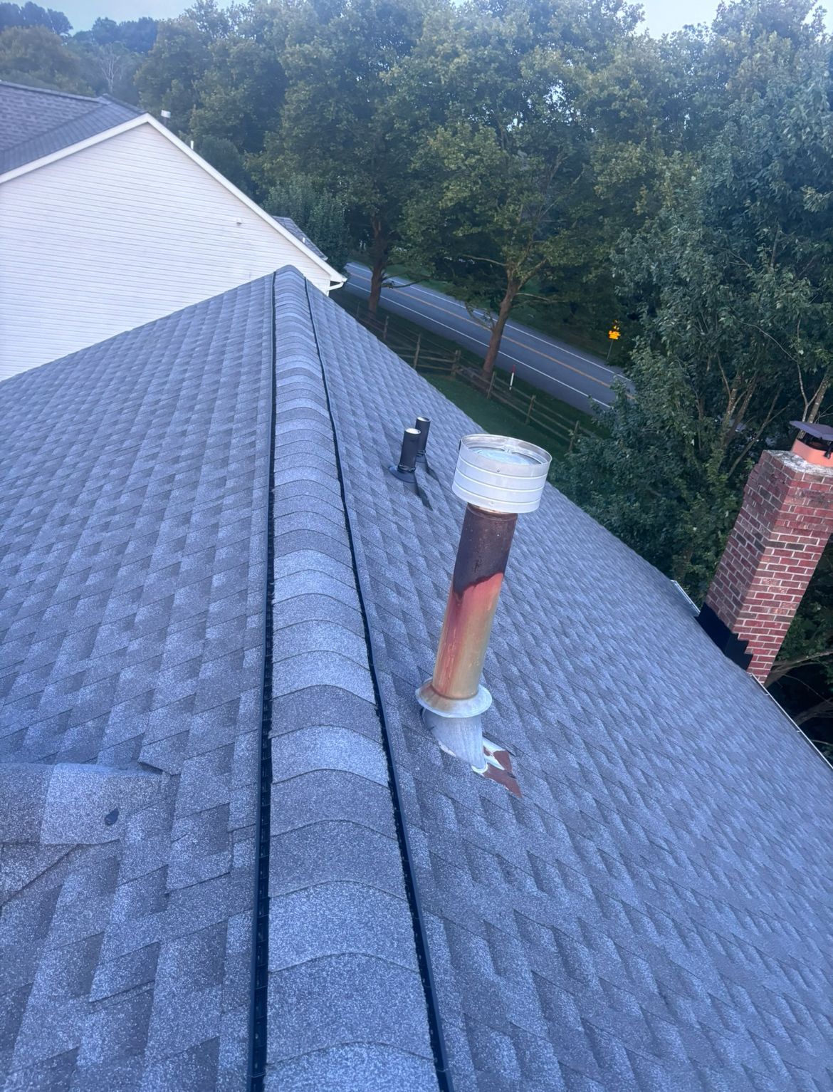Rooftop view of asphalt shingles, with a rusted vent pipe and a brick chimney in a residential setting.