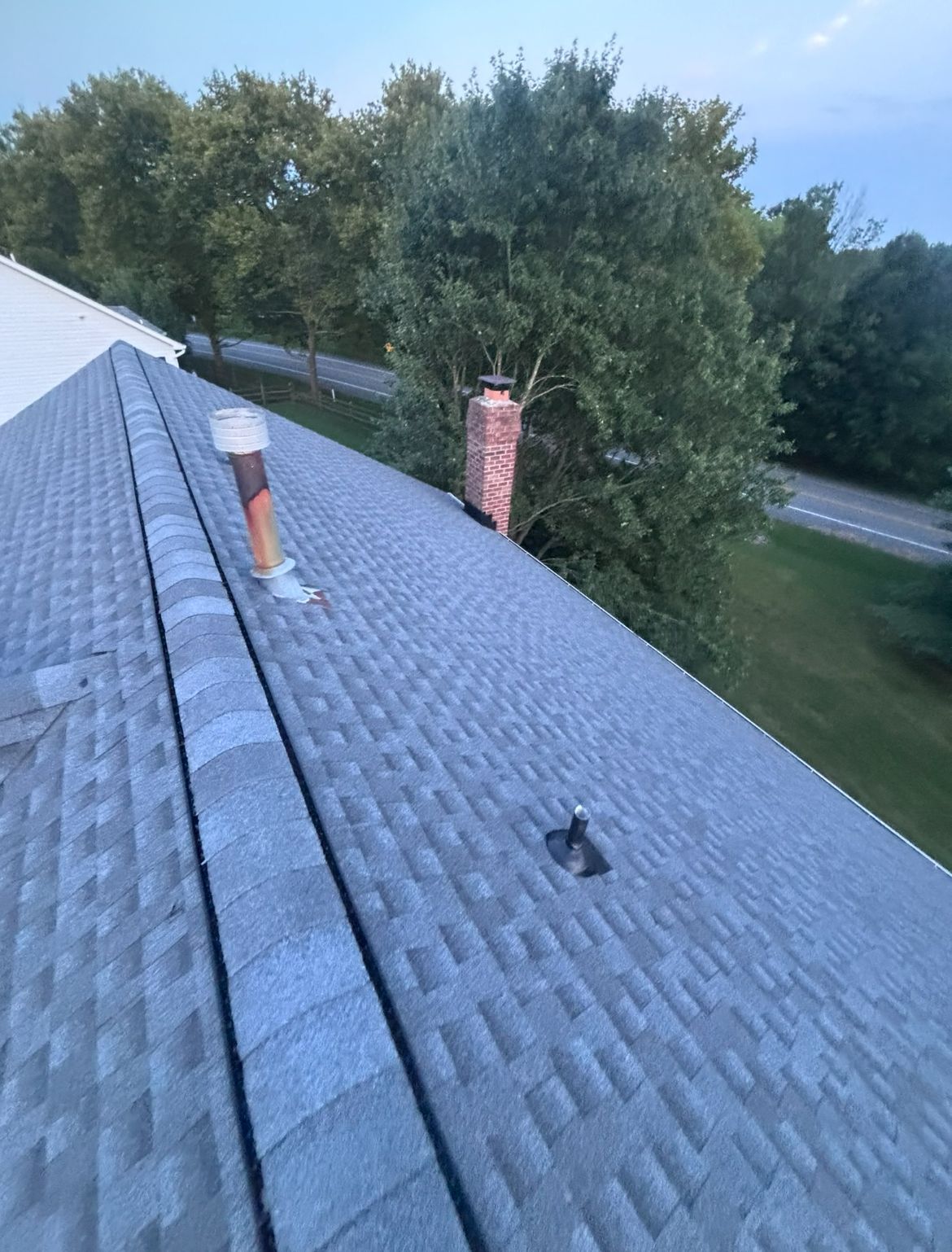 Gray shingled roof with a chimney and vent pipes against a backdrop of trees and a roadway.