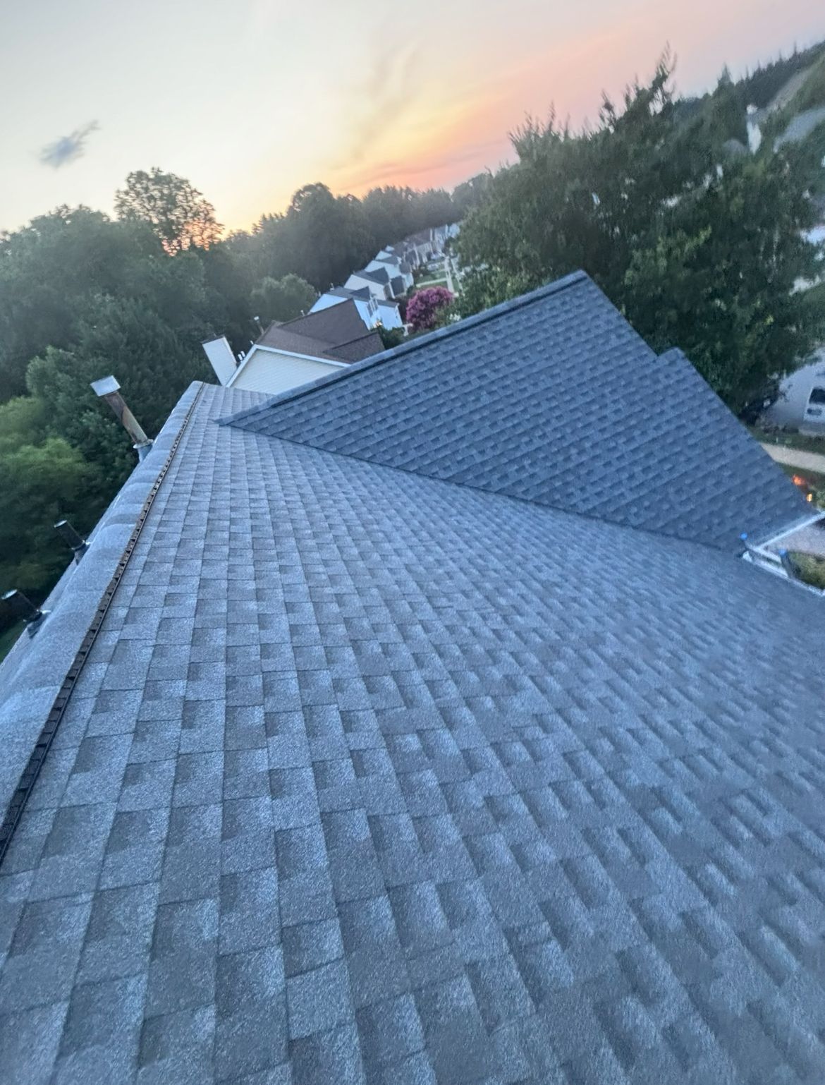 Gray asphalt shingle roof on a house at sunset, with neighborhood and trees in background.