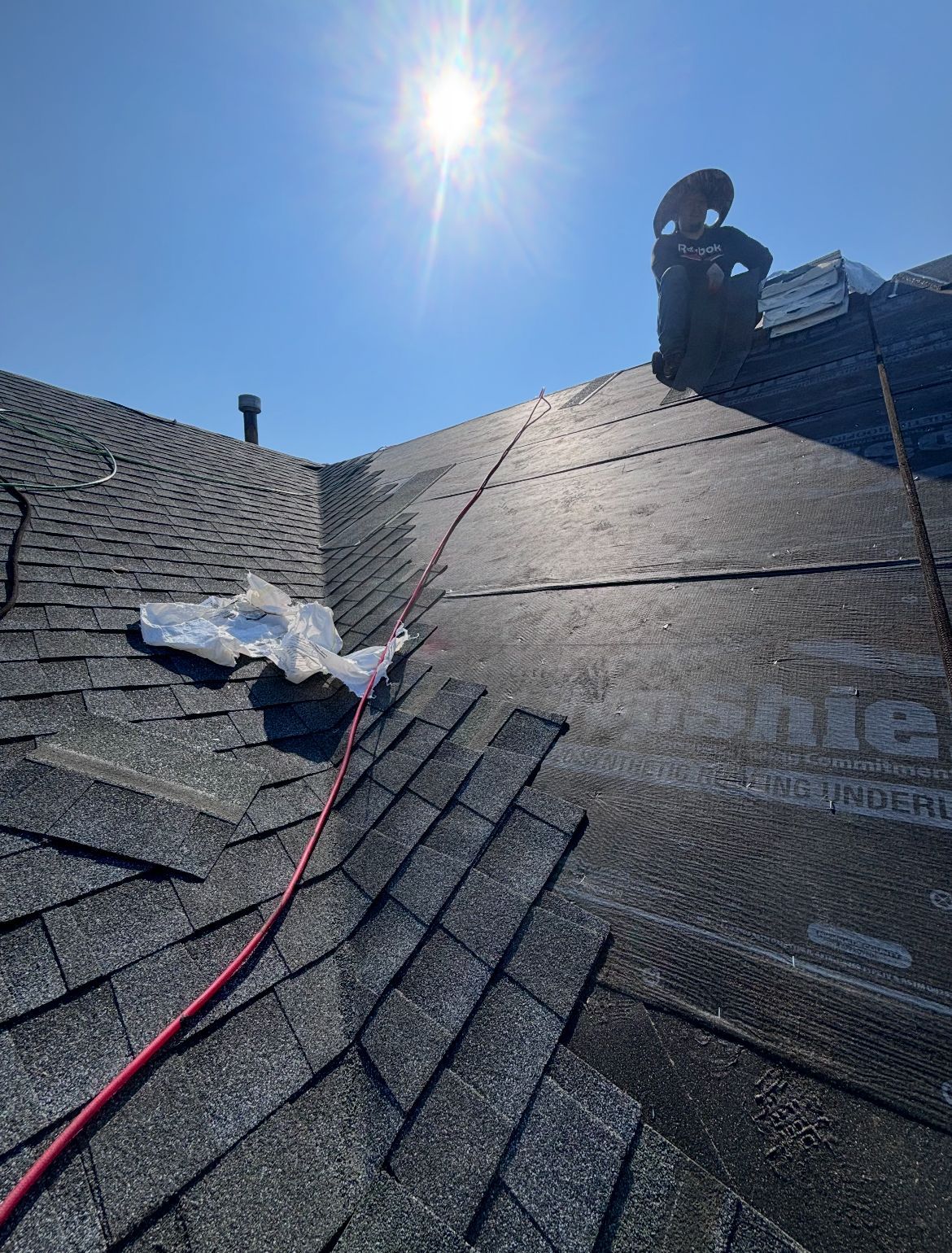 Roofer working on a shingled roof under a bright sun. Safety rope and chimney visible.