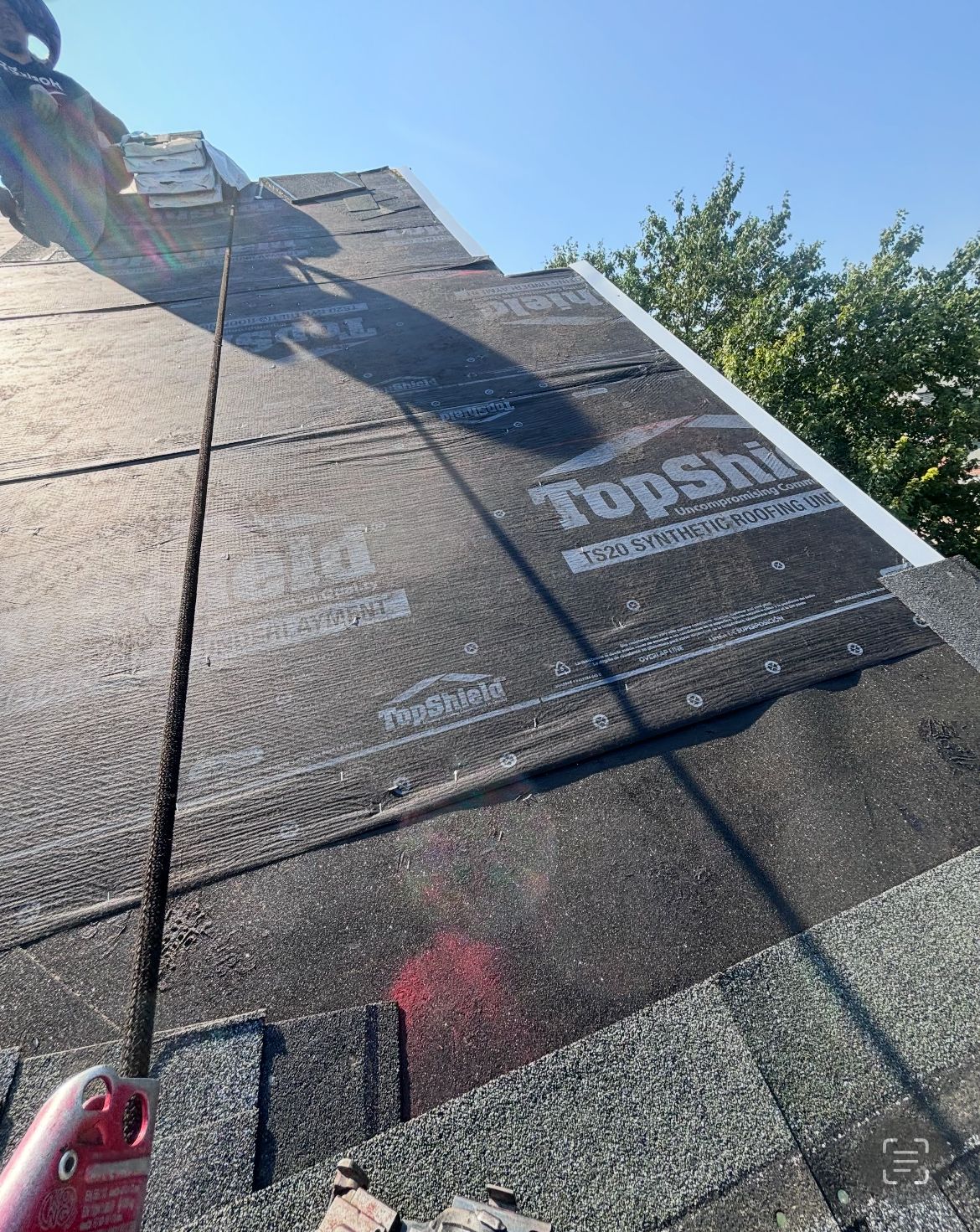 Roofer working on a roof, safety harness visible. Shingles are being replaced with some removed. Bright sunlight.