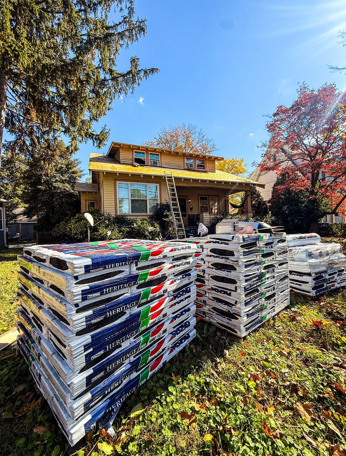 Stacks of roofing materials on a lawn with a house in the background being worked on under a blue sky.