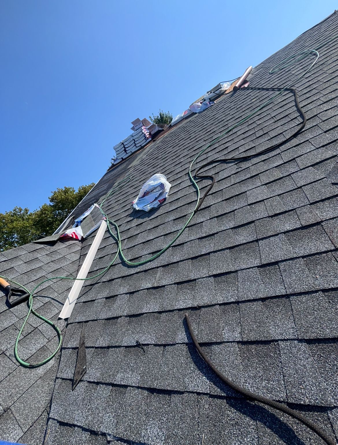 Rooftop with shingles under a clear blue sky. A hose, supplies, and tools are on the roof.