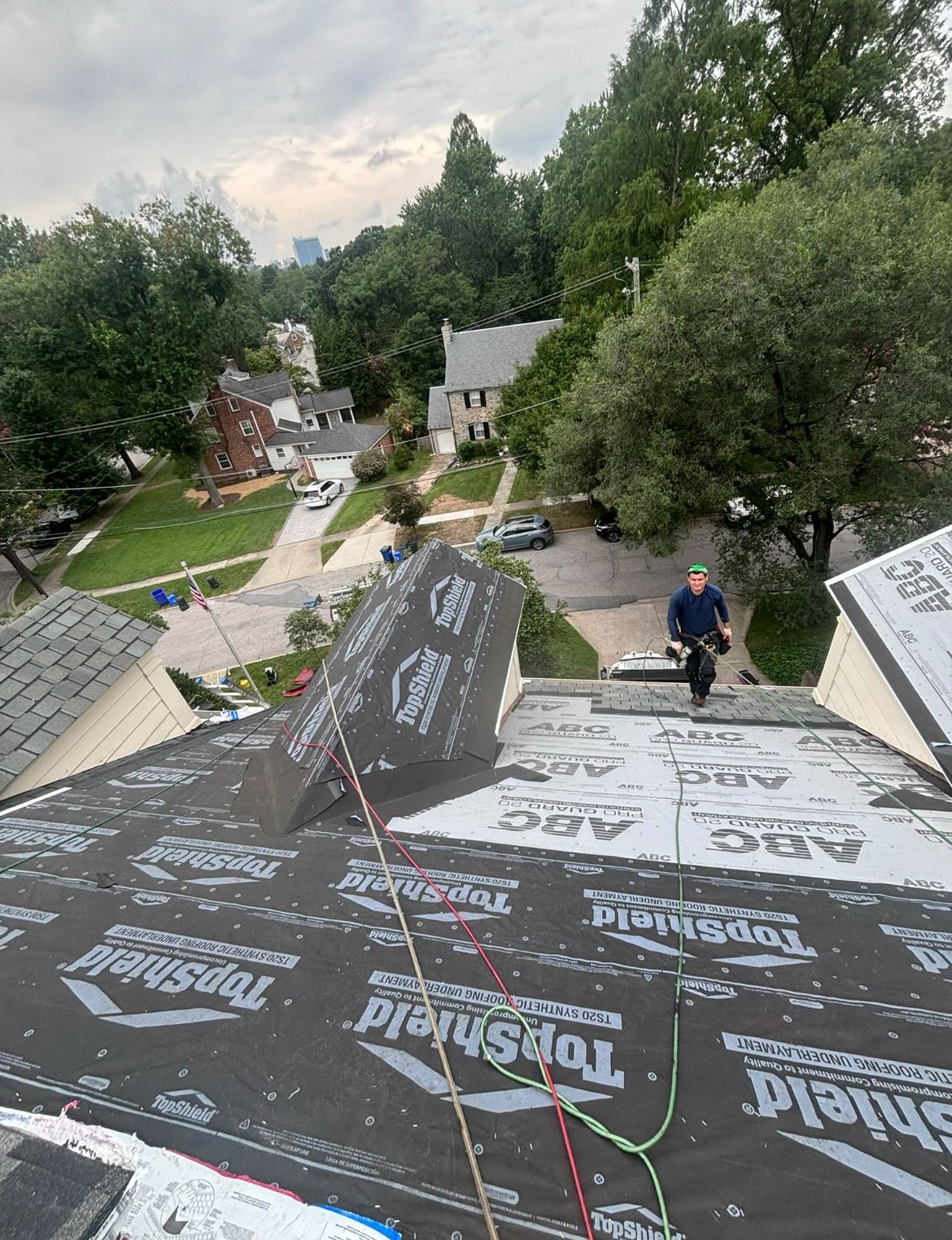 Roofer on a residential roof, installing roofing materials. Cloudy day, houses and trees in background.