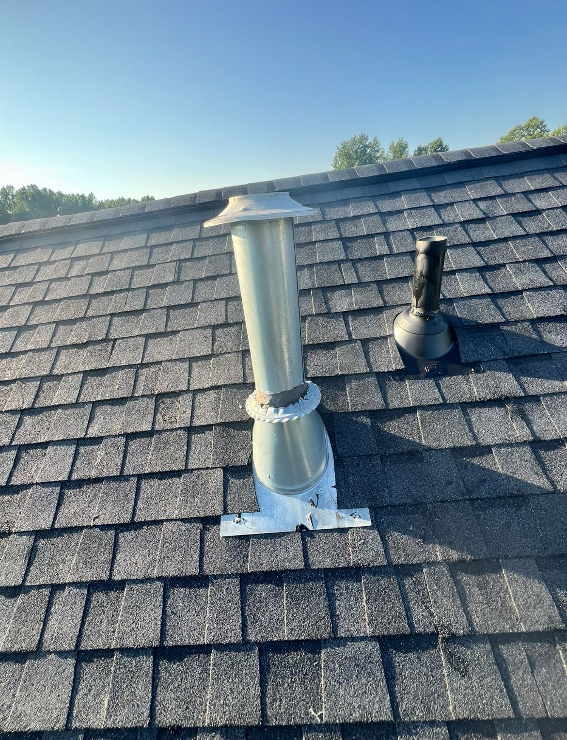 Two metal chimney vents on a gray shingle roof under a blue sky.