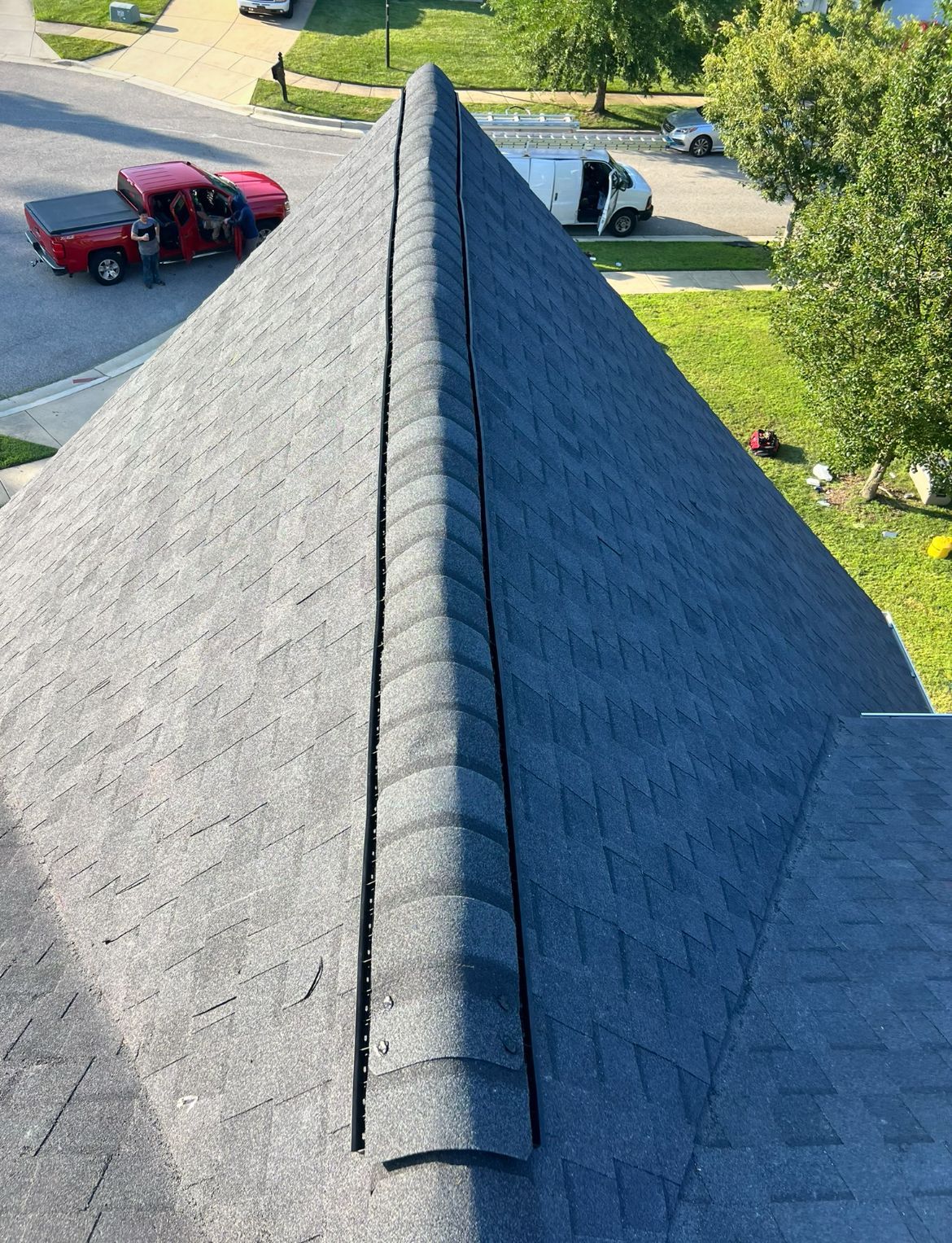 Overhead view of a dark gray asphalt shingle roof with a ridge cap. A red truck and van are in the background.