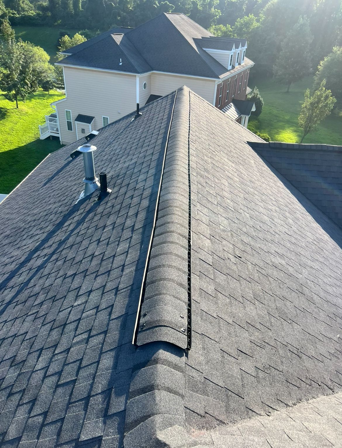 Dark asphalt shingle roof with a ridge vent. A chimney and large house are in the background.