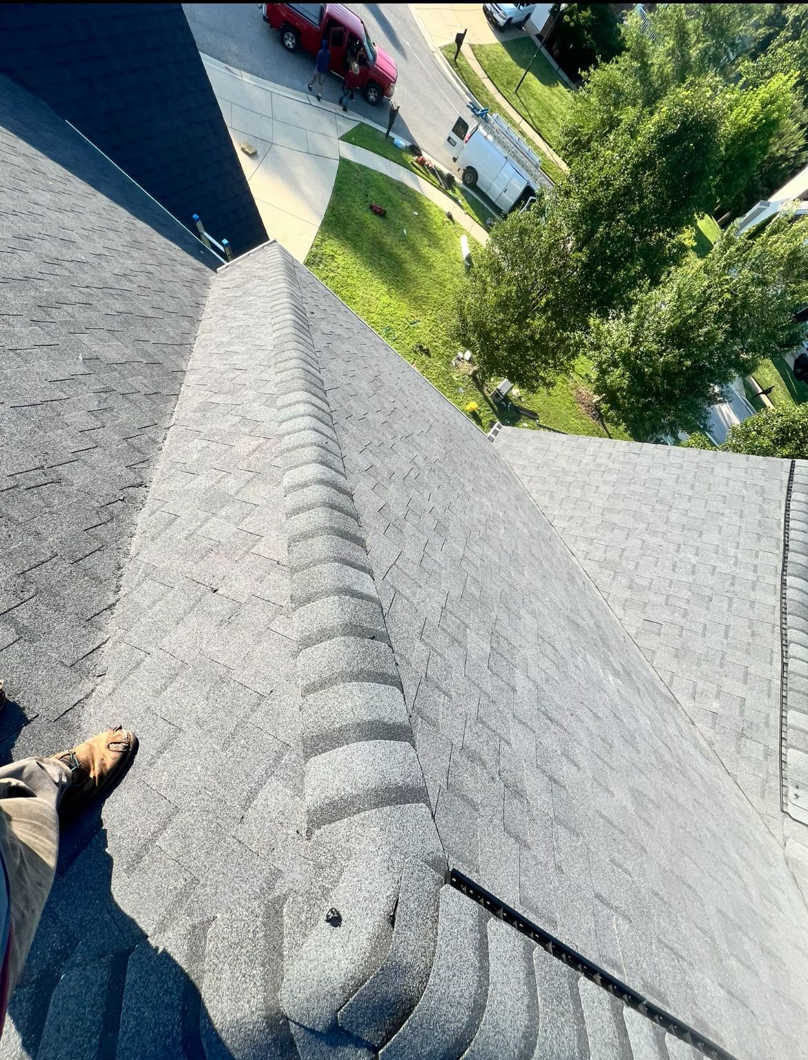 Looking down at a dark gray shingled roof with a ridgeline, view of a street and some trees.