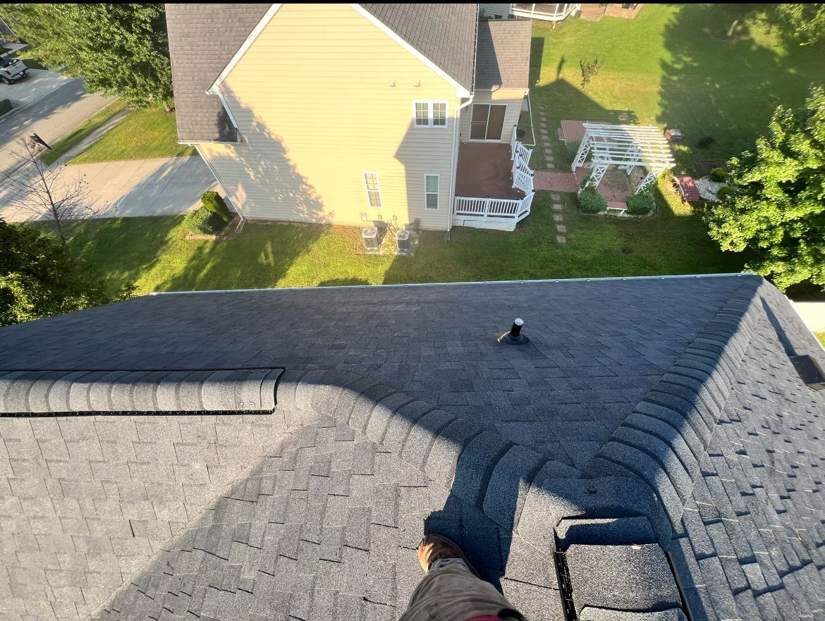 A person's feet on a dark roof, looking down at a house and lawn.