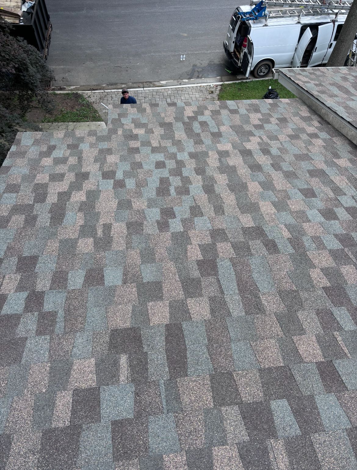 Overhead view of a roof with a checkerboard pattern of brown, gray, and blue shingles, near a street with parked vehicles.