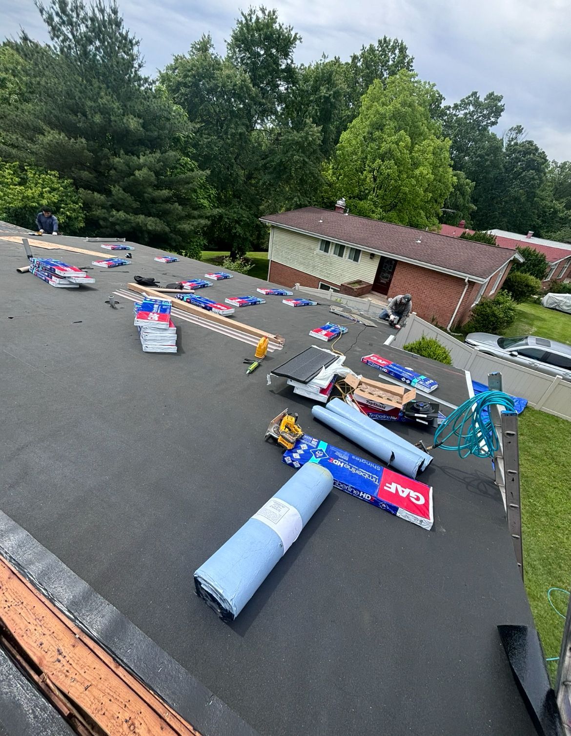 Rooftop with roofing materials: rolls, bundles, and tools. House and trees in the background.