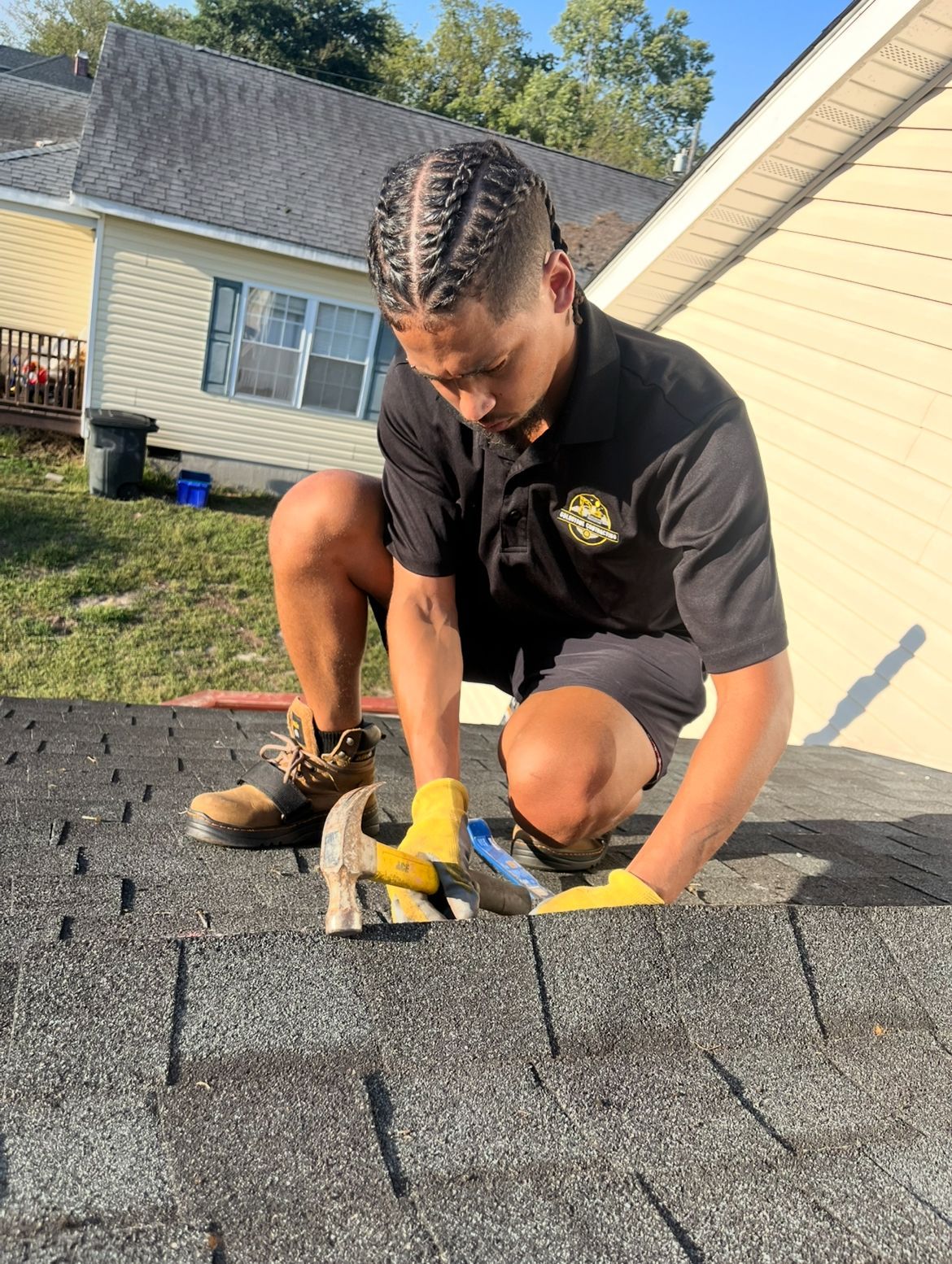 Person kneeling on a roof, hammering shingles. Wearing gloves and work boots. Sunny outdoor setting.