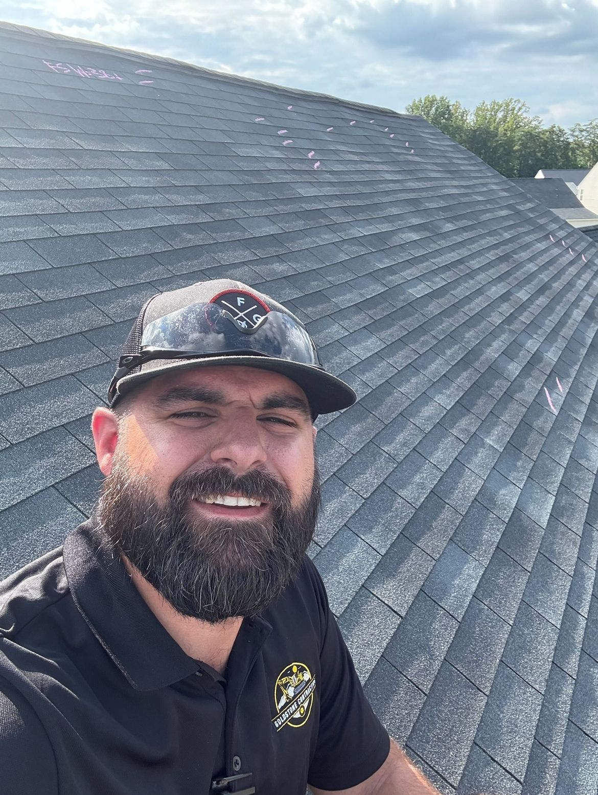 Man wearing a hat smiles on a roof. Asphalt shingles, cloudy sky.