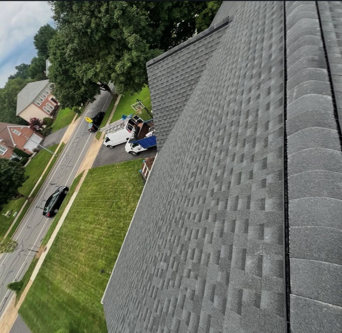 View from a rooftop: grey shingle roof, green grass, road with cars, houses, and trees.