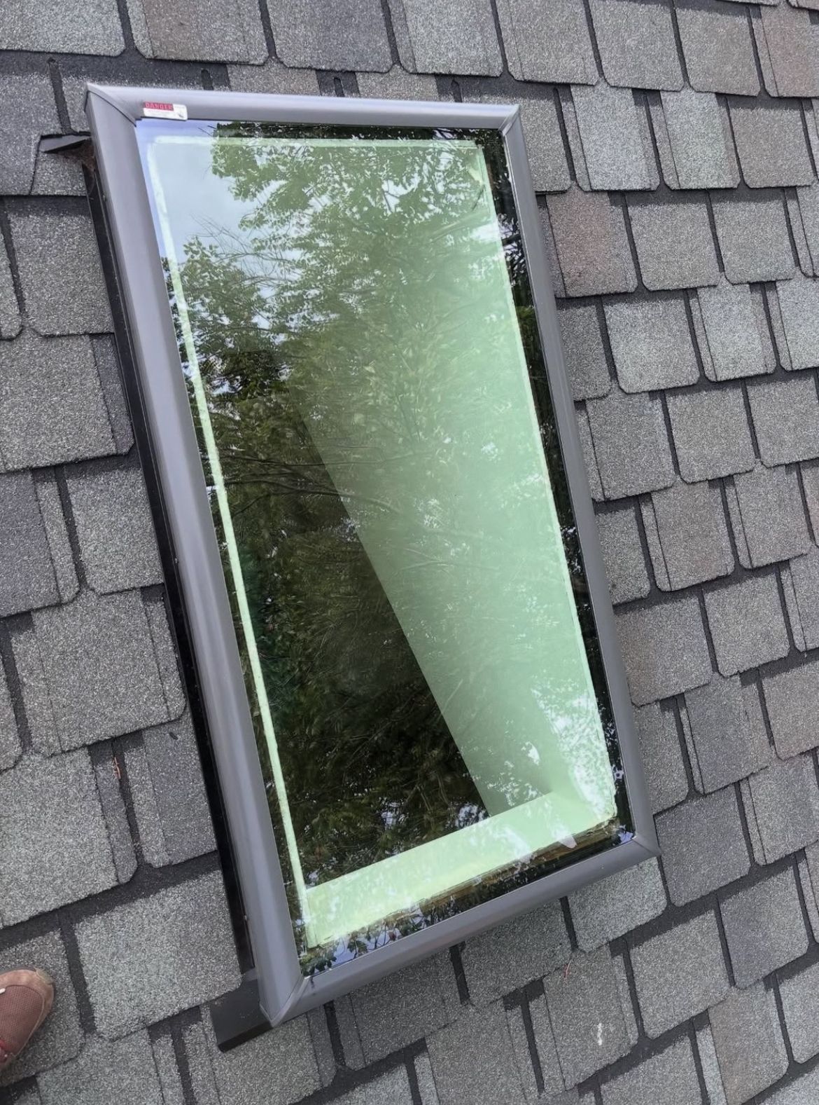 Skylight on a gray shingled roof, showing a reflection of trees and a metallic frame.