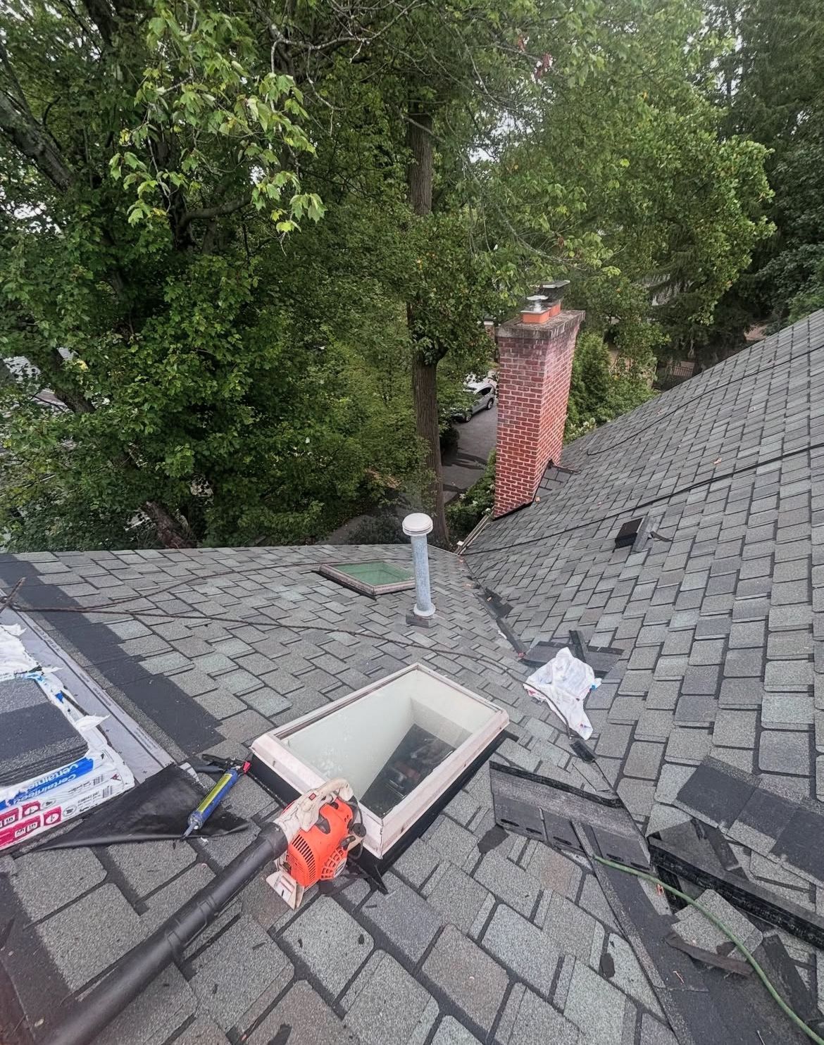 Rooftop with skylight, chimney, and tools; gray shingles; surrounded by green trees.