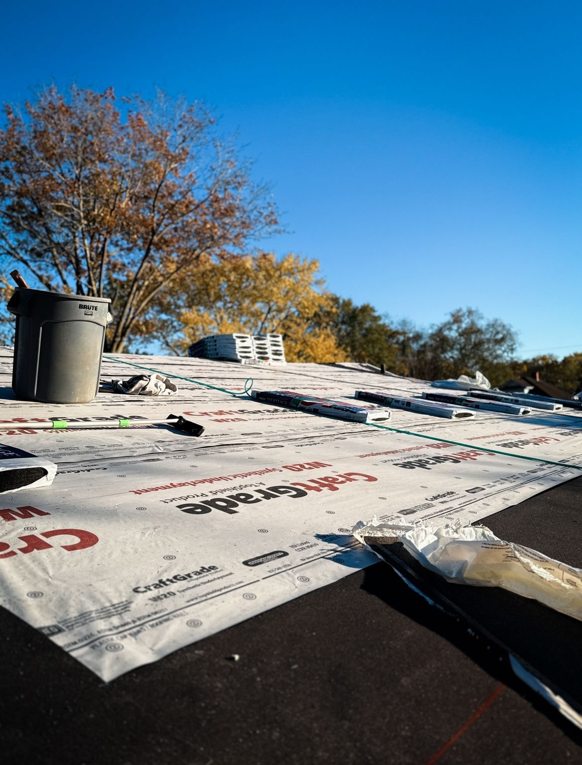 Rooftop with construction materials and partial white underlayment, blue sky, trees in the background.