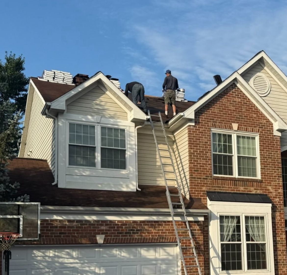 Roofers on a house roof installing shingles. A ladder leans against the roof. Sunny day.