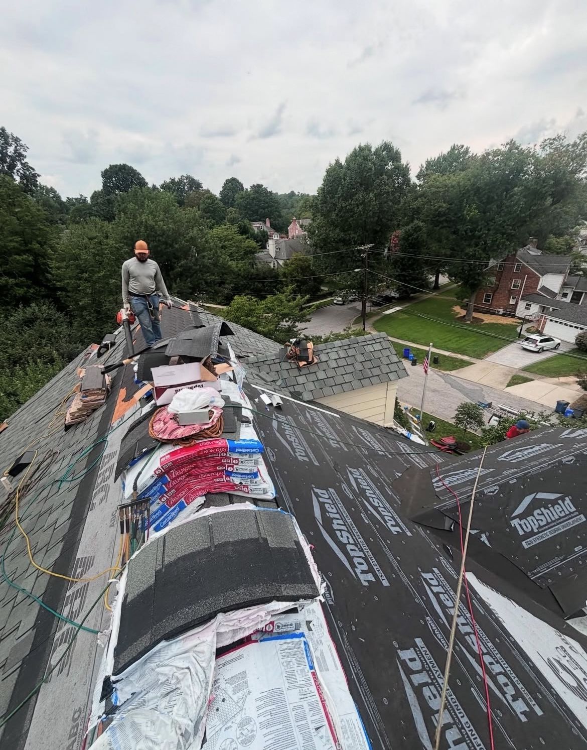Man on roof replacing shingles, overlooking neighborhood on cloudy day.