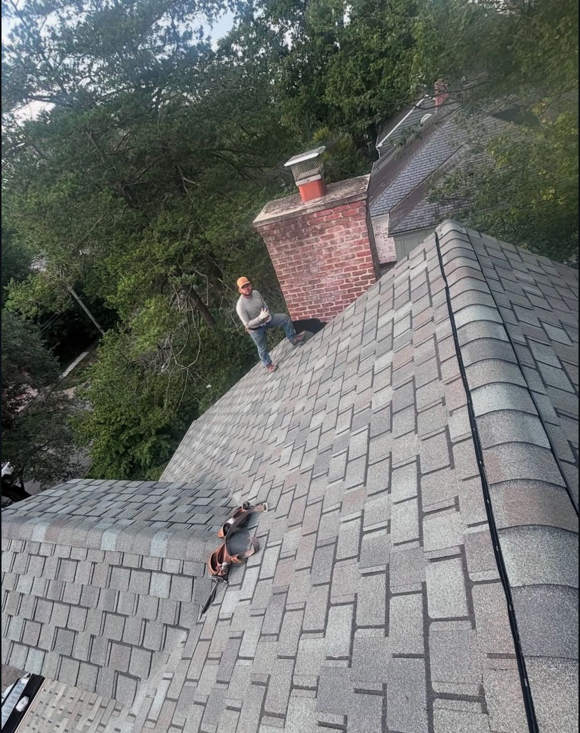 Person on a gray shingle roof near a brick chimney. Trees surround the house.