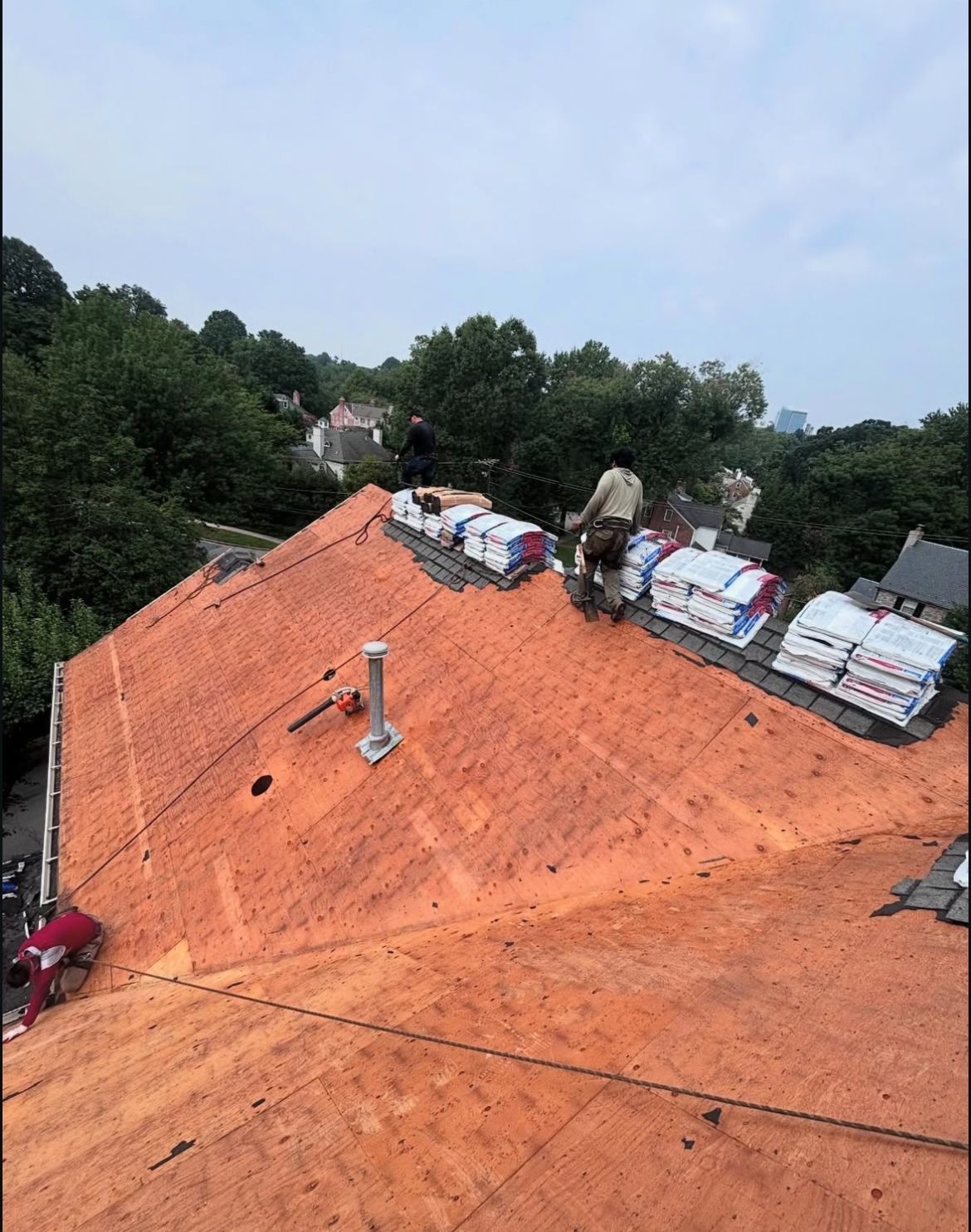 Roofers working on a house roof, surrounded by trees. The roof has exposed sheathing with bundles of shingles.