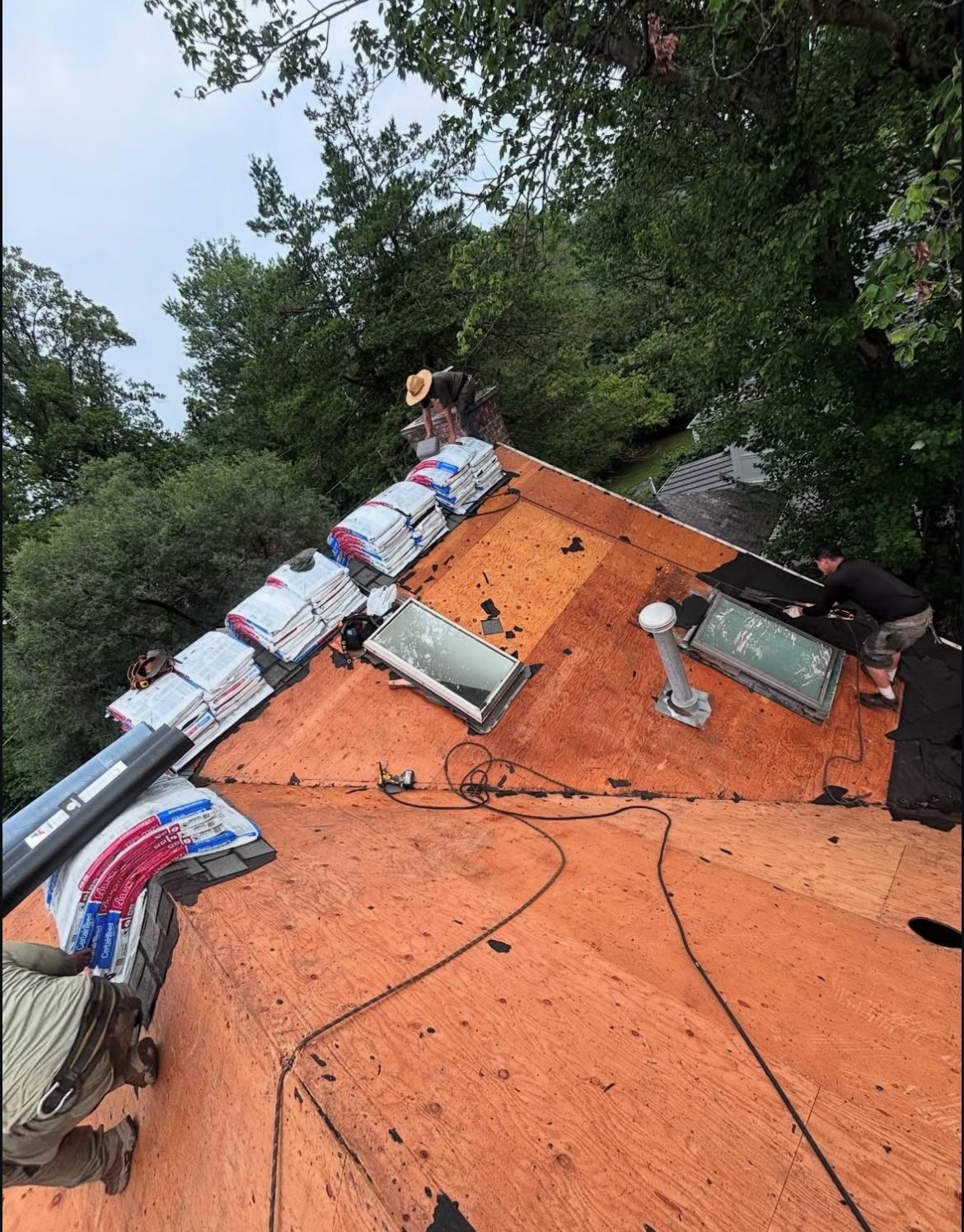 Roofers working on a roof, with shingles and skylights visible, surrounded by trees.