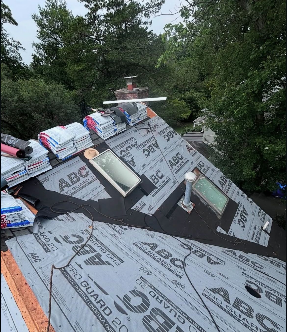 Rooftop under construction with skylights. Gray underlayment and shingles with stacks of materials.