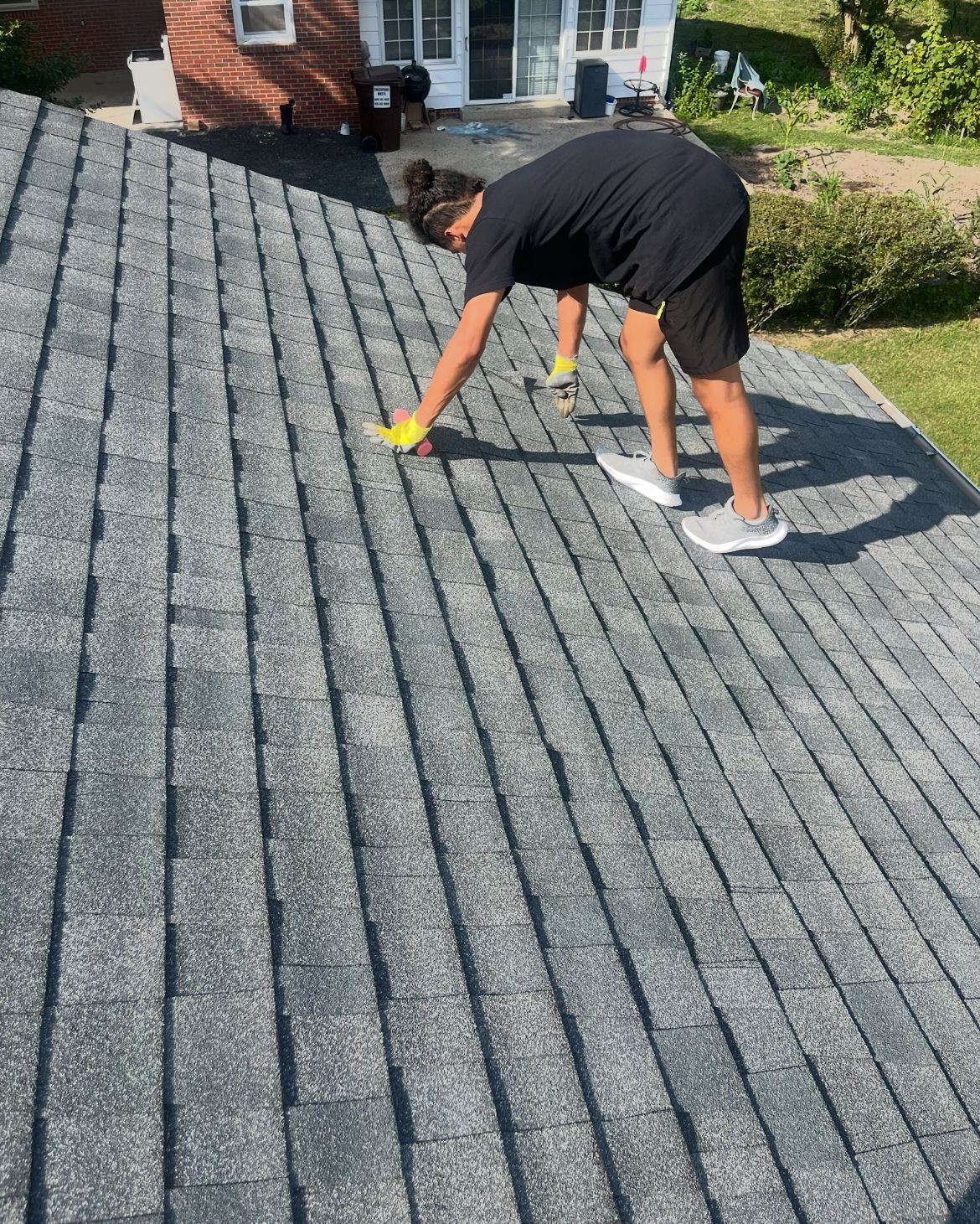 Person on a roof inspecting gray shingles, sunny day.
