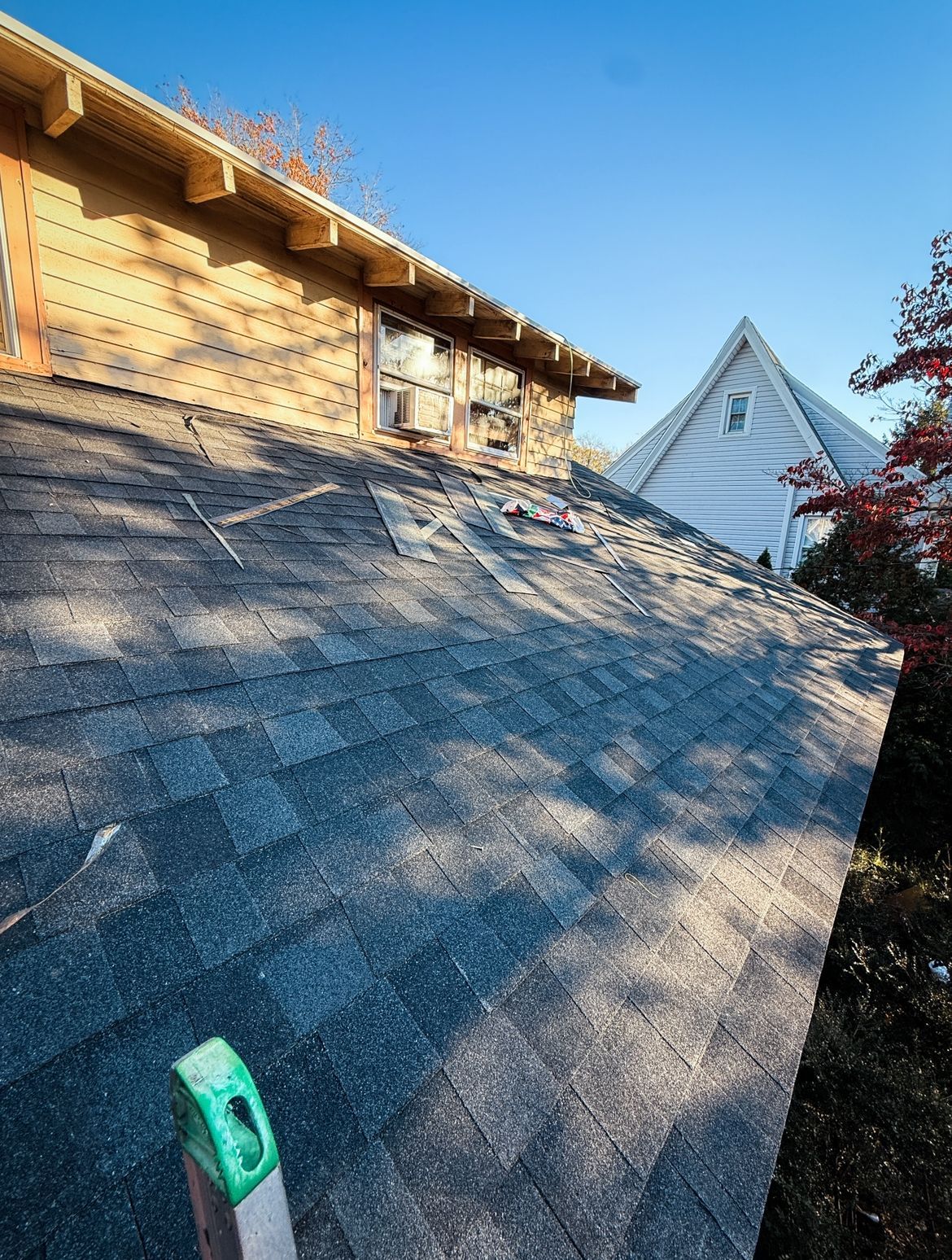 Shingled roof with visible damage; a nearby house and clear sky are also in view.