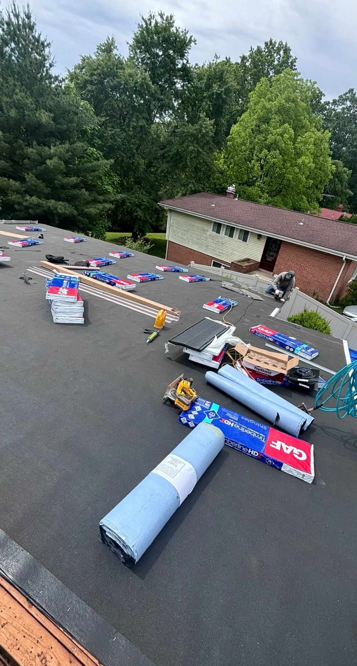 Roofing materials scattered on a flat roof with a house and trees in the background.