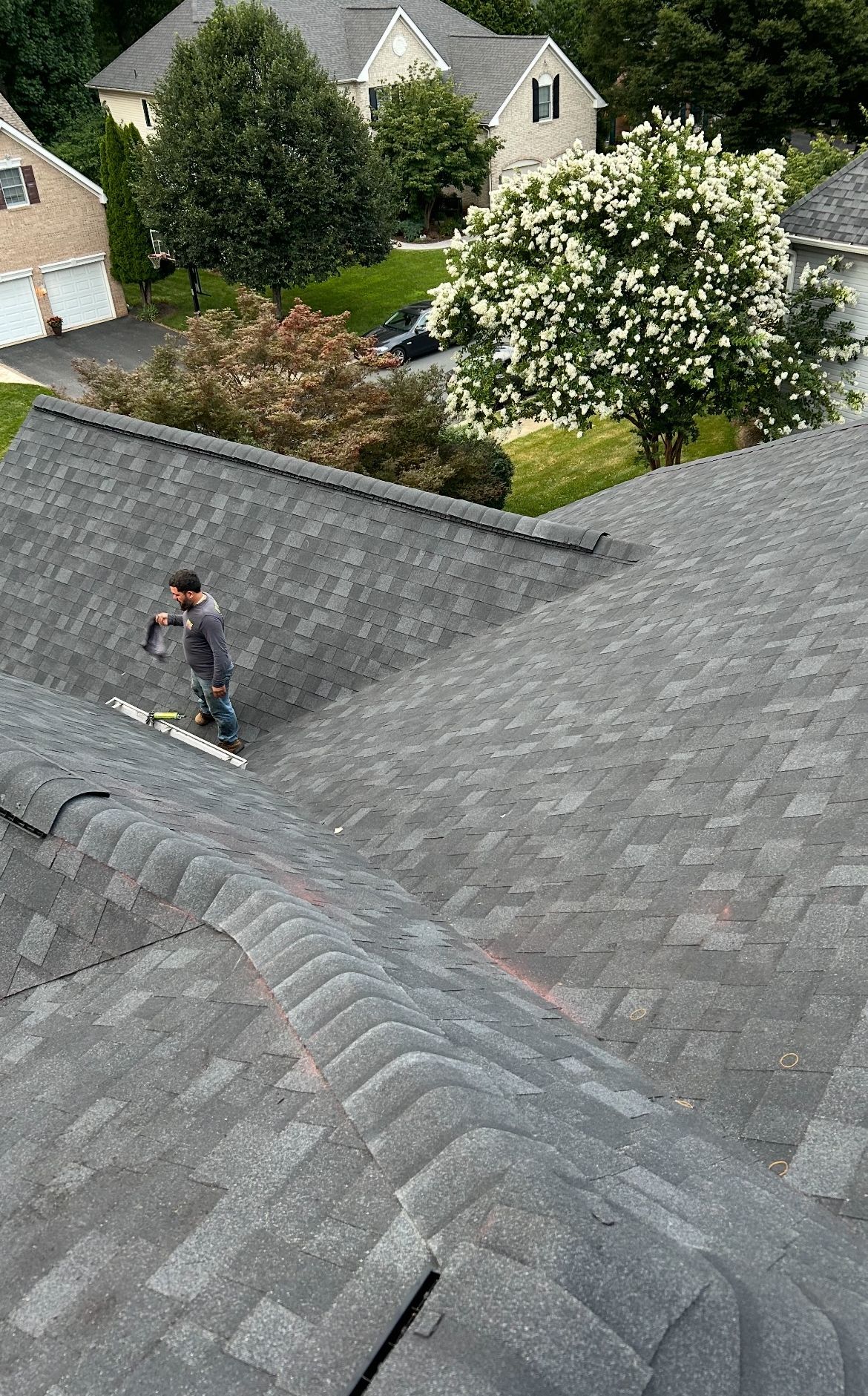 Person on a roof, repairing shingles on a house. Other houses and trees are visible in the background.