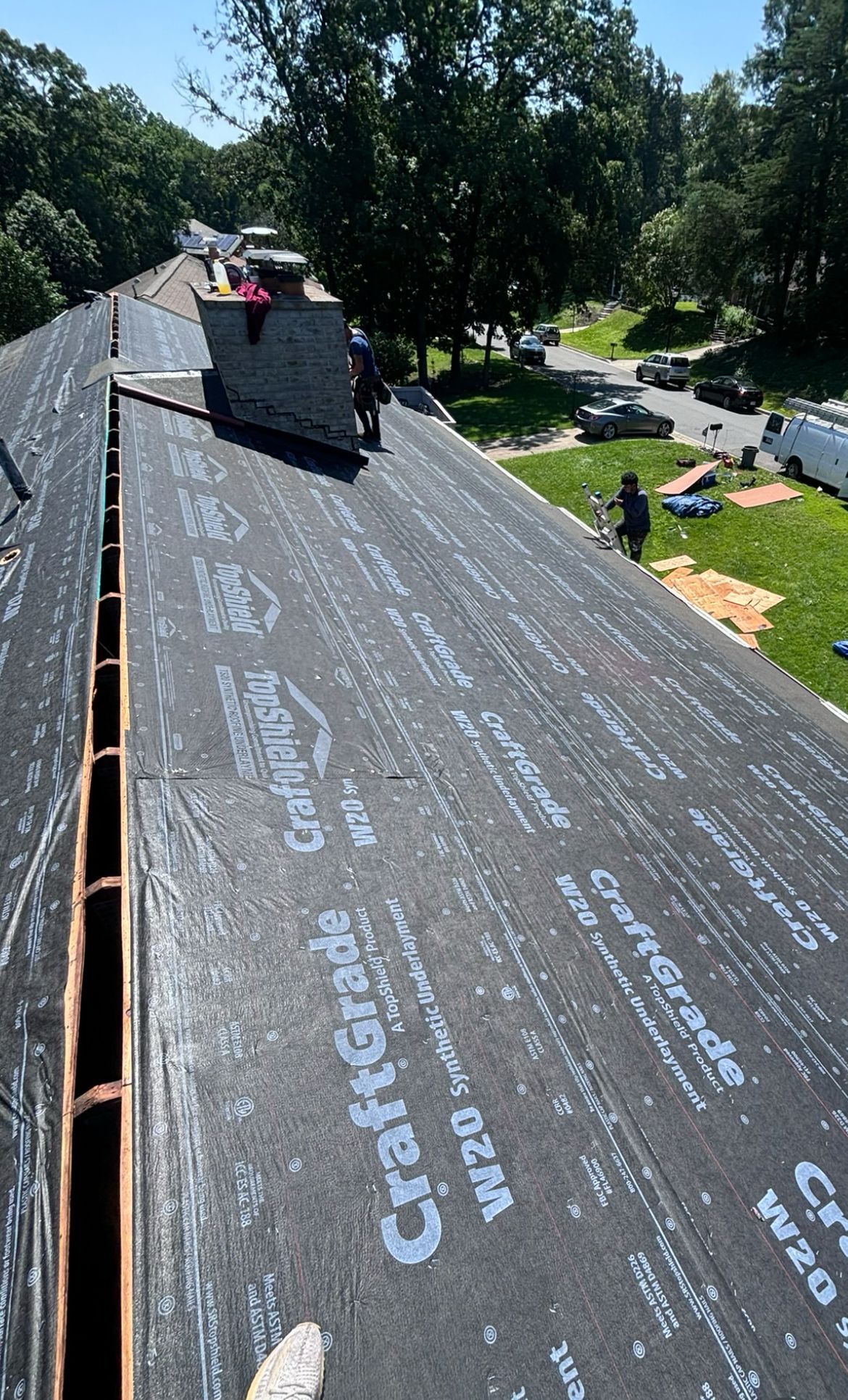 Roofers installing roofing material on a residential roof, with green trees and parked cars in the background.
