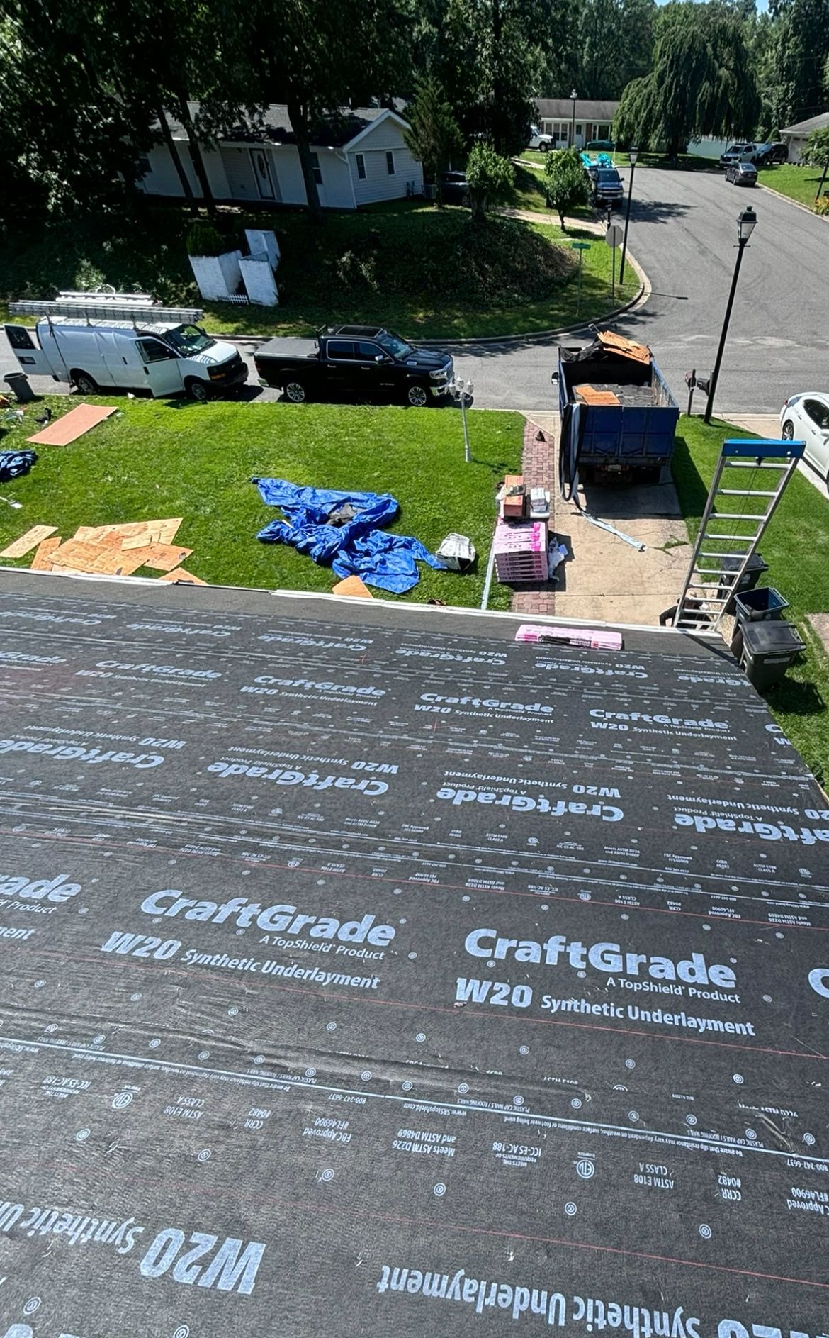Roofer installing roofing materials, with a view of a street, trees, and parked vehicles.