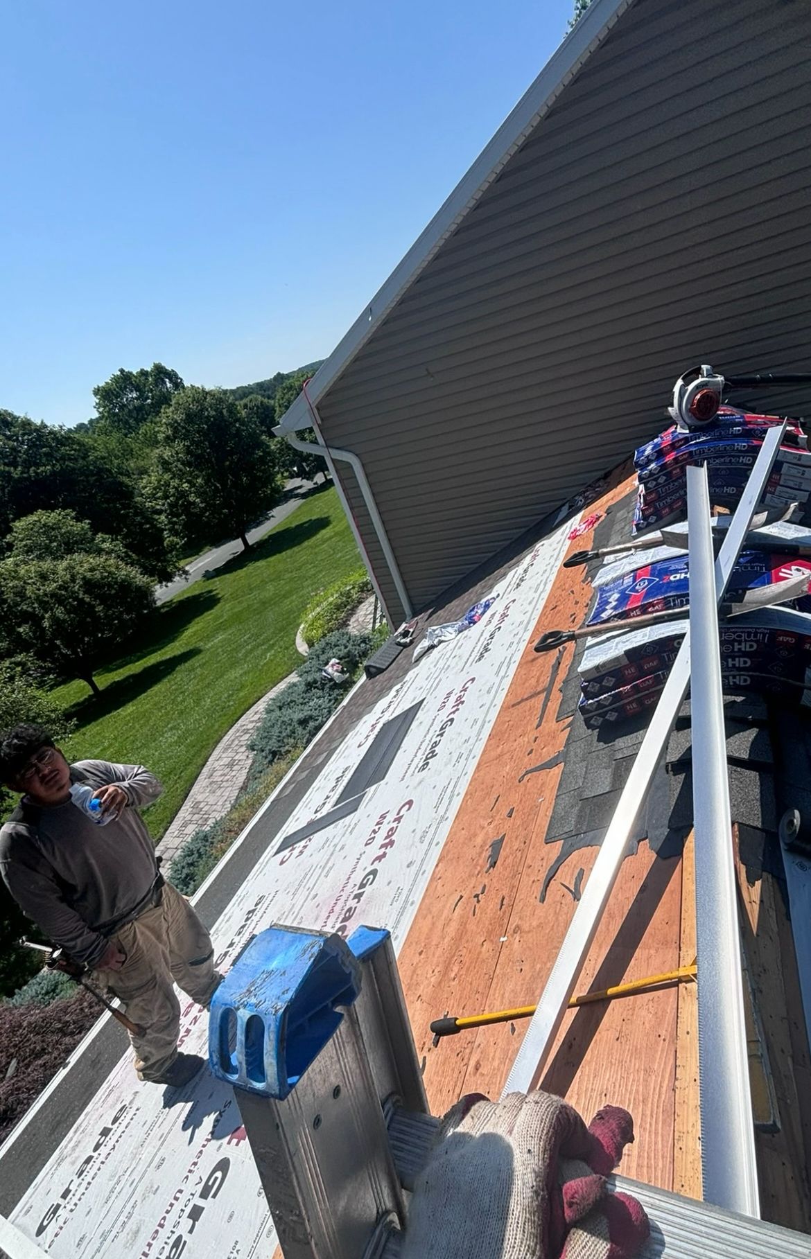 Workers on a roof removing shingles, ladder propped, blue sky.