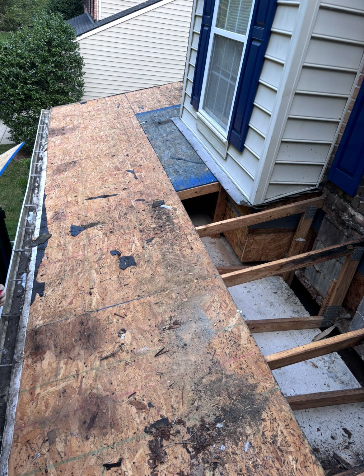 Damaged roof with exposed sheathing, near a window with blue shutters, exterior view.