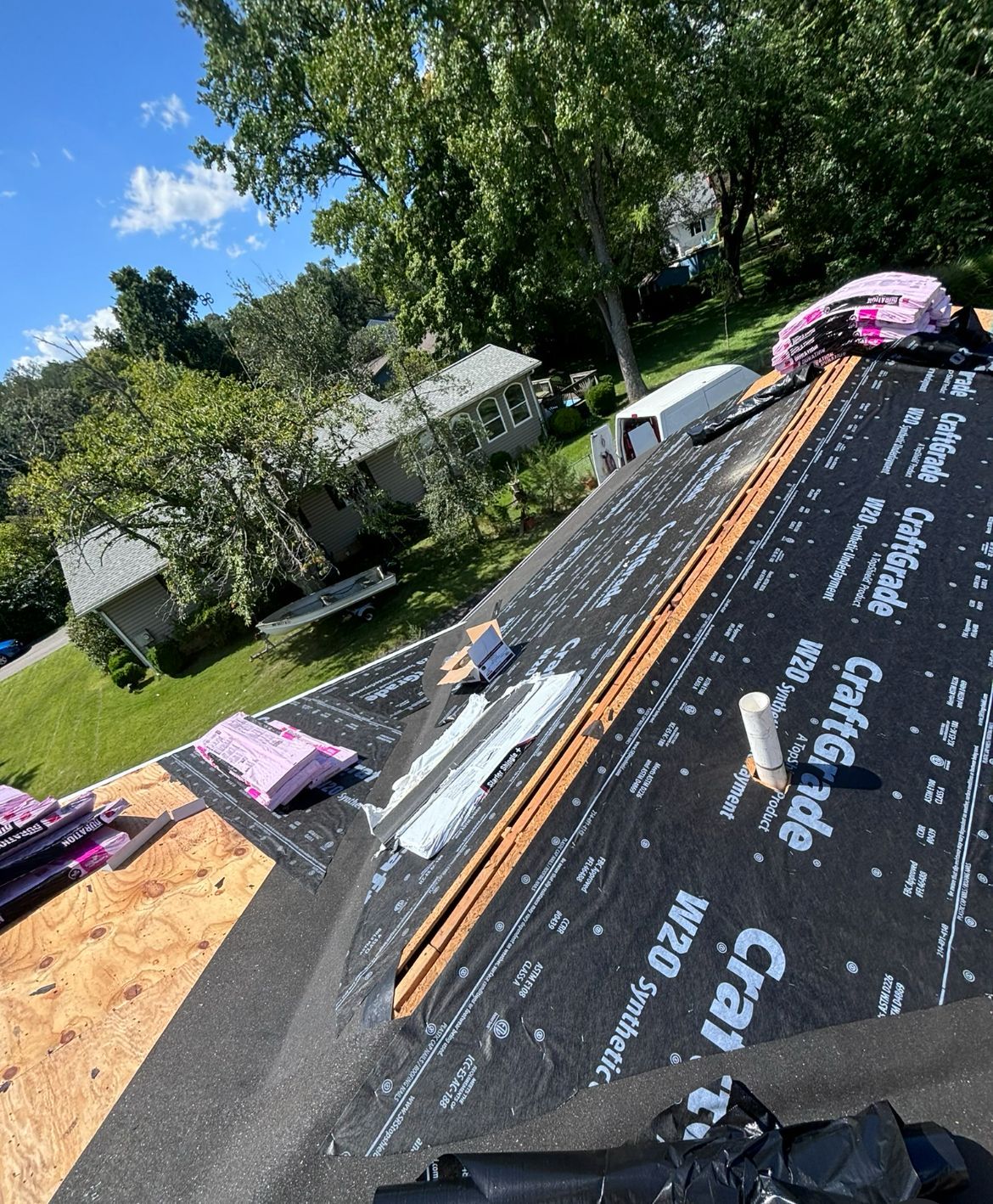 Rooftop with black underlayment, pink insulation, and bundles of shingles; sunny day, trees in background.