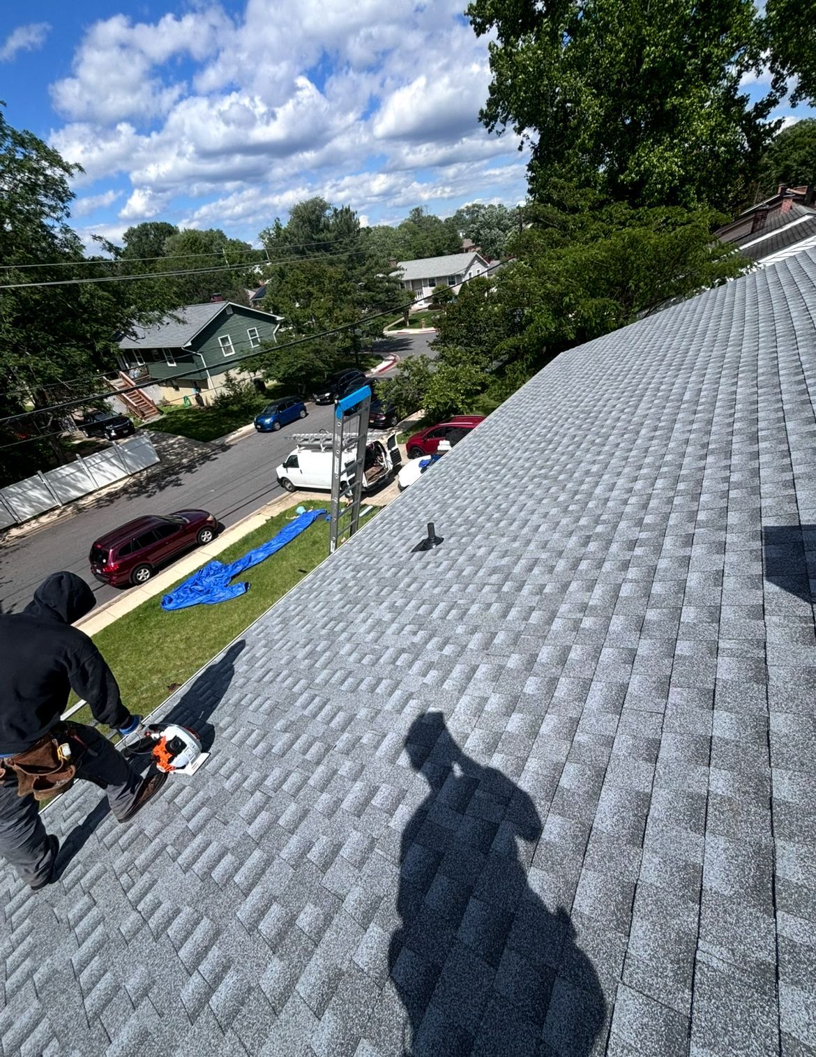 Roofer on gray shingle roof, cutting shingles with a saw on a sunny day; neighborhood in the background.