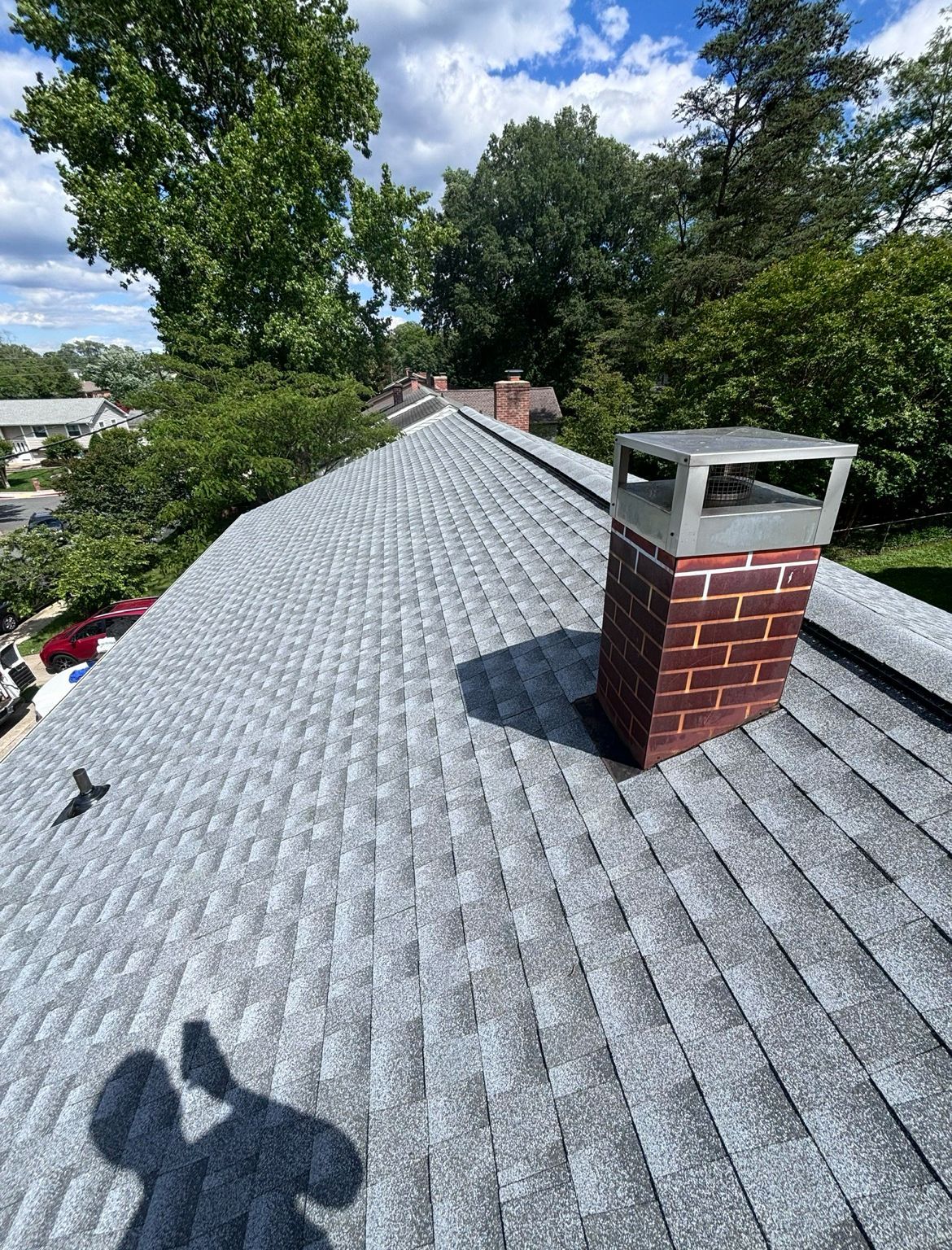 Gray shingled roof with a brick chimney and metal cap against a blue sky, trees in background.