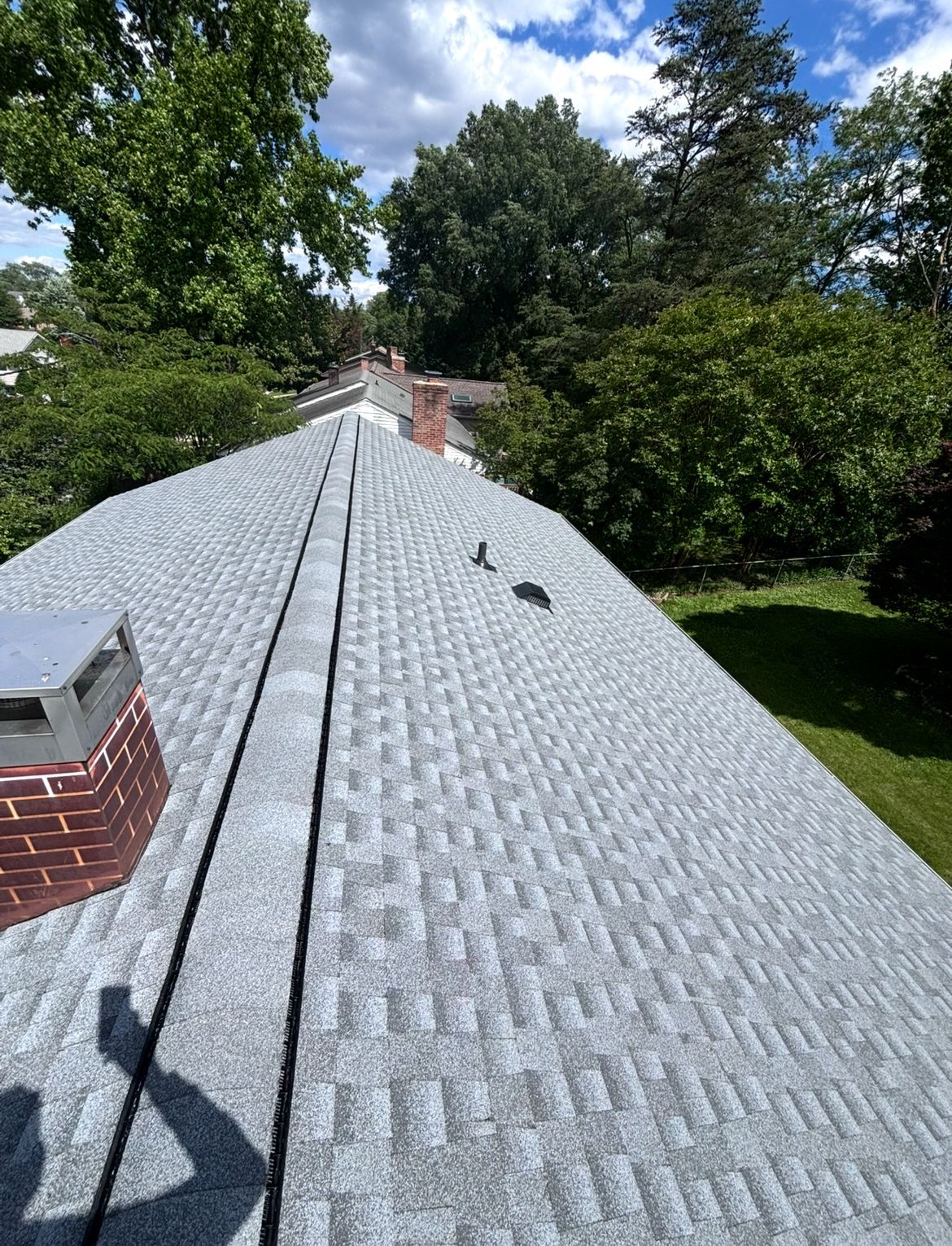 Gray asphalt shingle roof with a brick chimney and lush green trees under a blue sky.